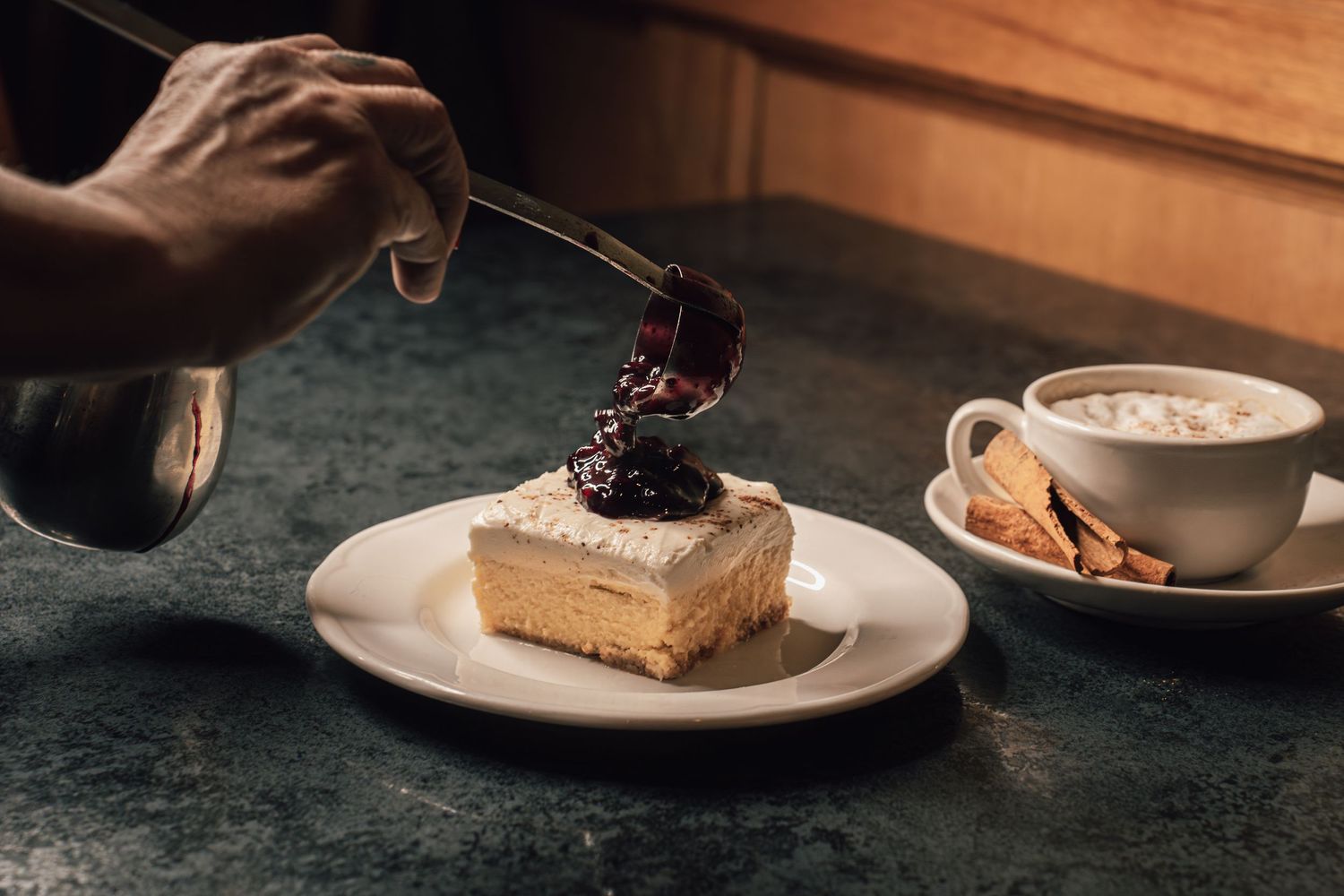Dark berry sauce being poured over a creamy layered cake slice next to a cappuccino cup.