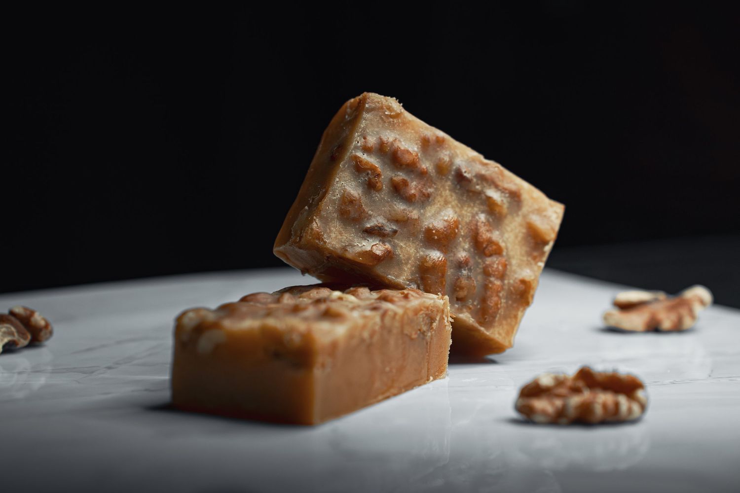 Two pieces of walnut fudge candy on a marble surface with whole walnuts, dark background.