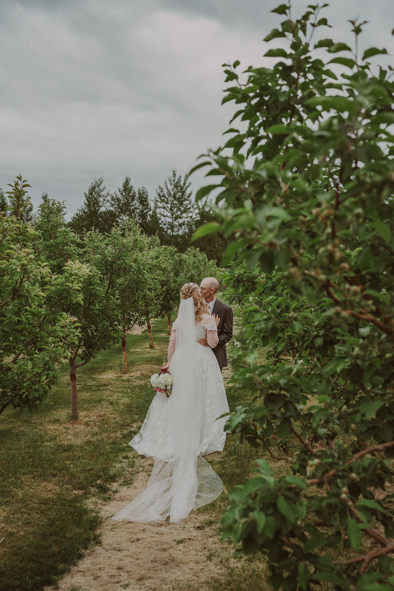 Bride and groom kissing on sandy path through green orchard under cloudy sky at Northern Lights Winery, Prince George, BC.
