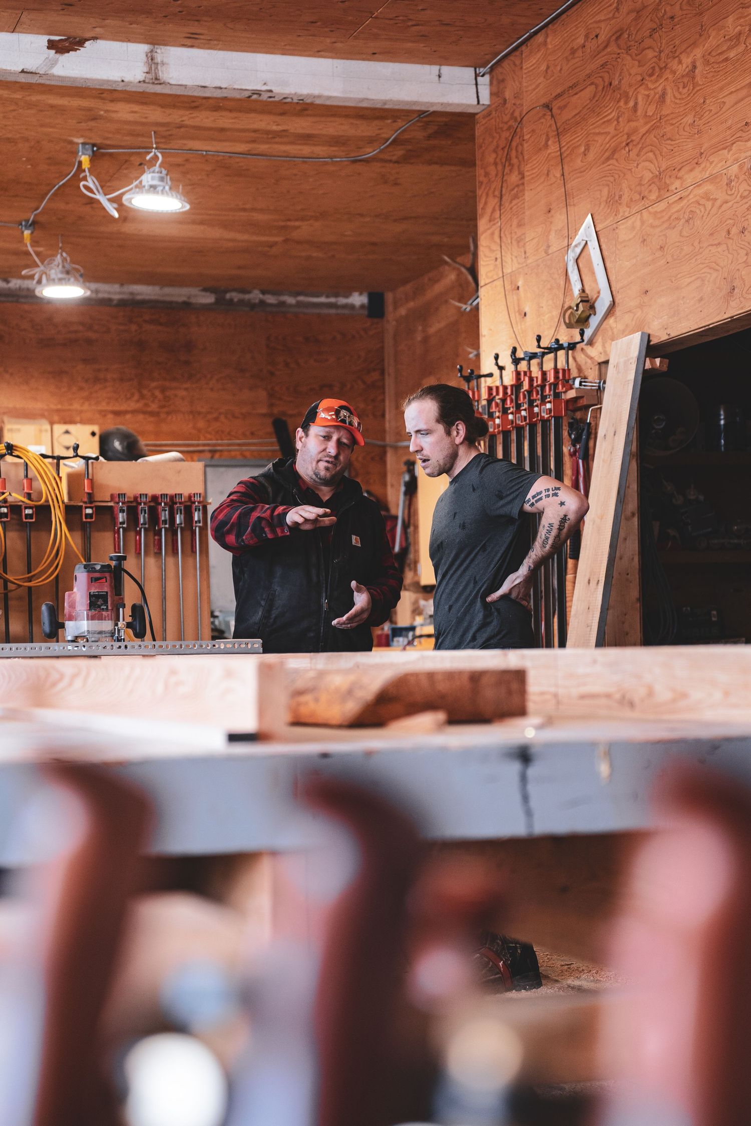 Two men in a woodworking workshop discussing a project surrounded by tools and lumber.
