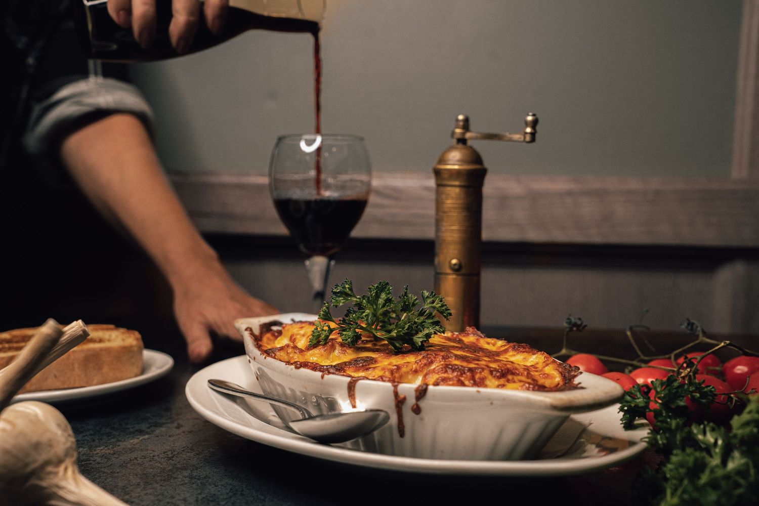 Server presenting a golden-topped savory pie beside a glass of red wine and pepper grinder at a restaurant.