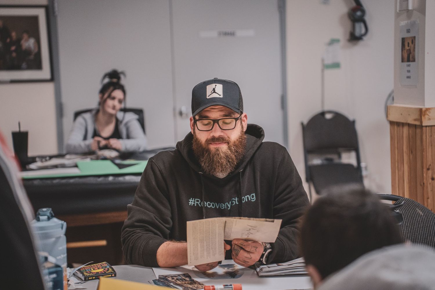 Bearded man in a Jordan cap and hoodie smiles while holding papers at an indoor community workshop table.