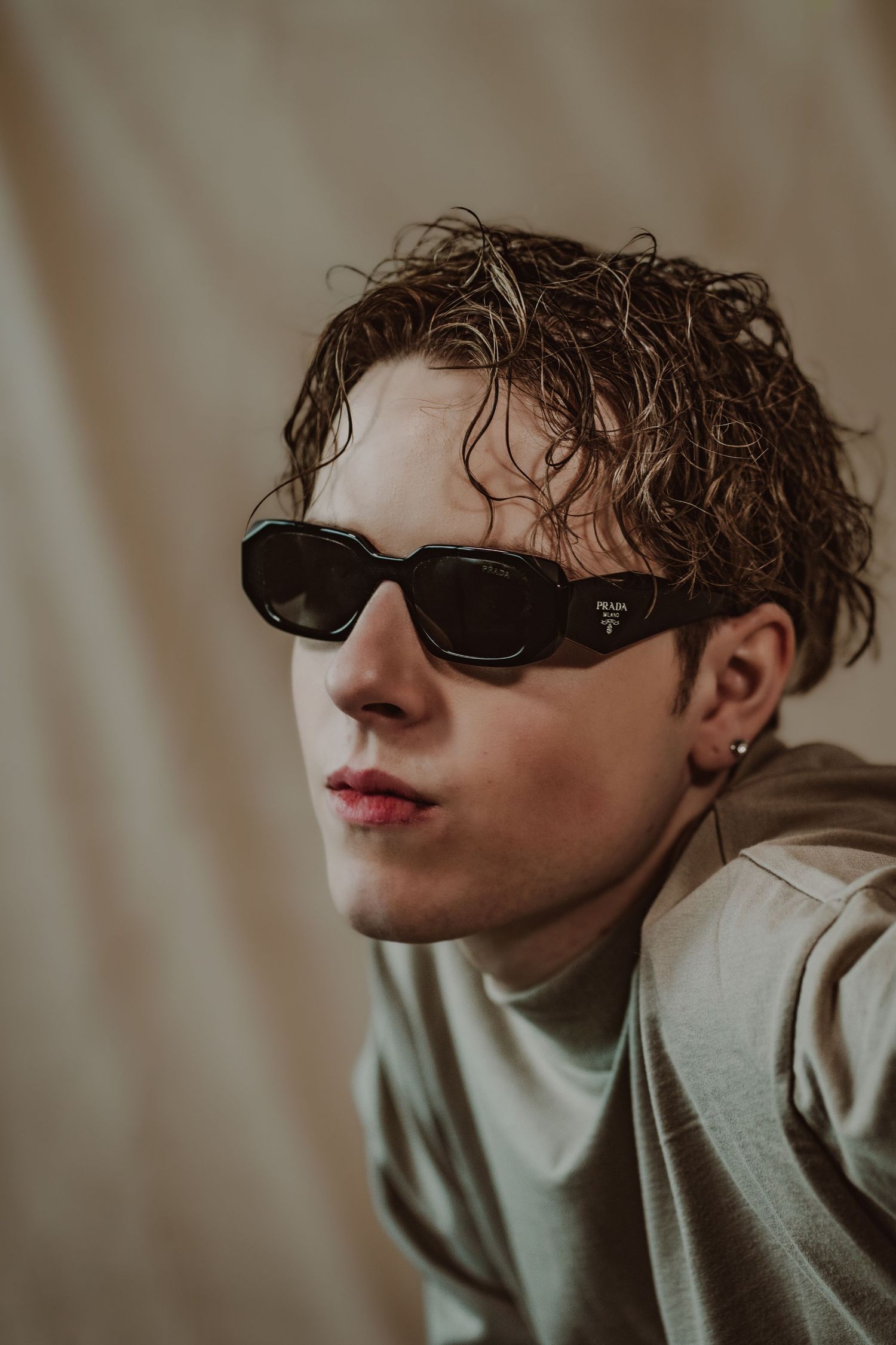Young man with curly wet hair wearing Prada black sunglasses and gray hoodie against beige background during model photoshoot.