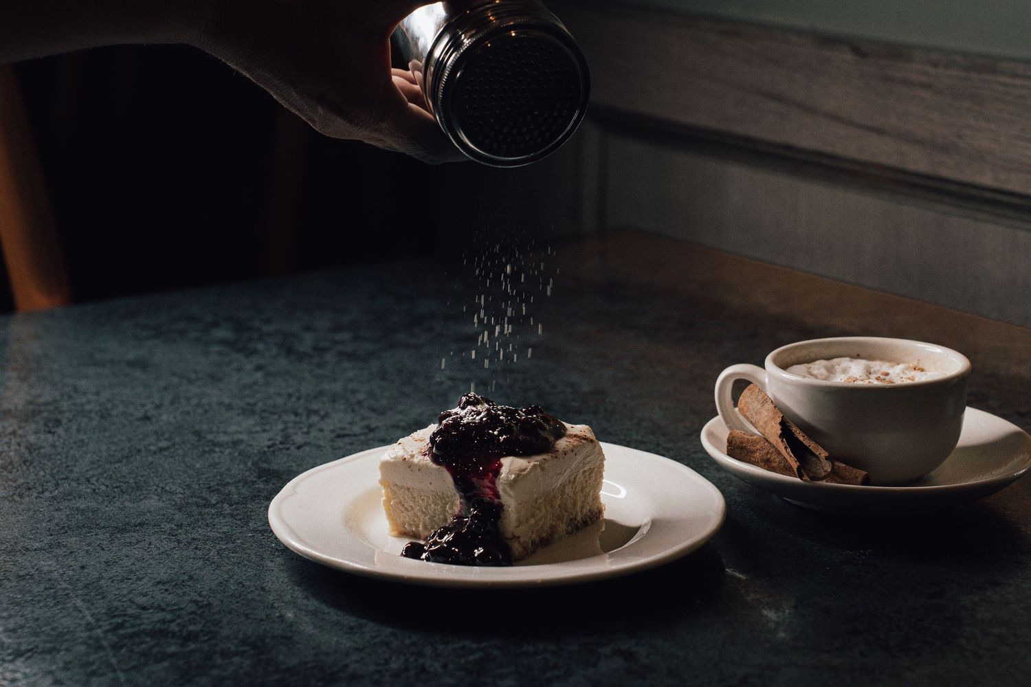 Powdered sugar being dusted over a blueberry cheesecake slice beside a cappuccino on marble table.