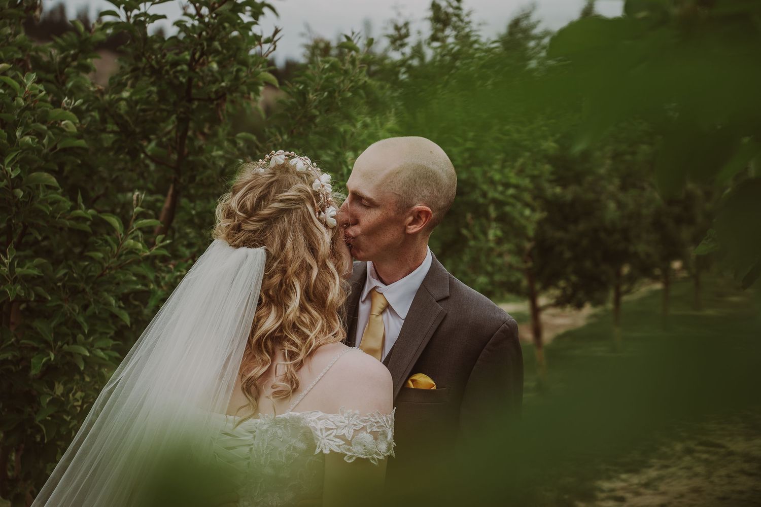 Bride and groom sharing a kiss surrounded by lush green trees in outdoor orchard setting at Northern Lights Winery, Prince George, BC.