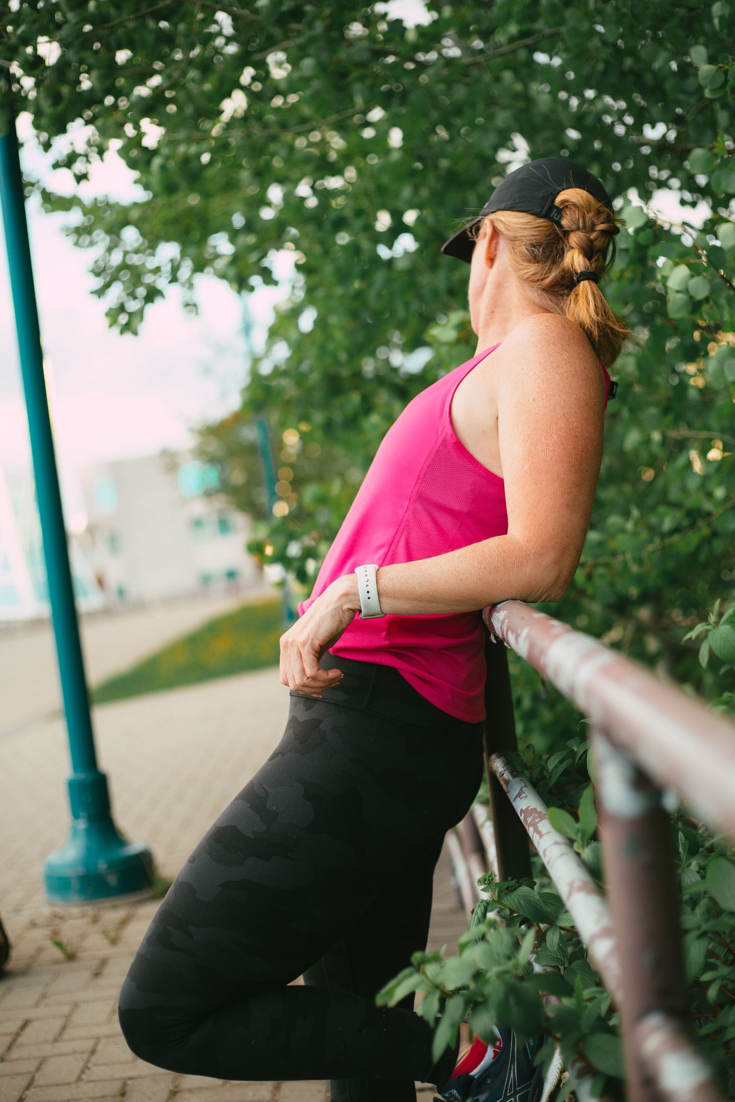 A woman in a pink tank top and black leggings leans against a railing surrounded by green trees during commercial photoshoot.