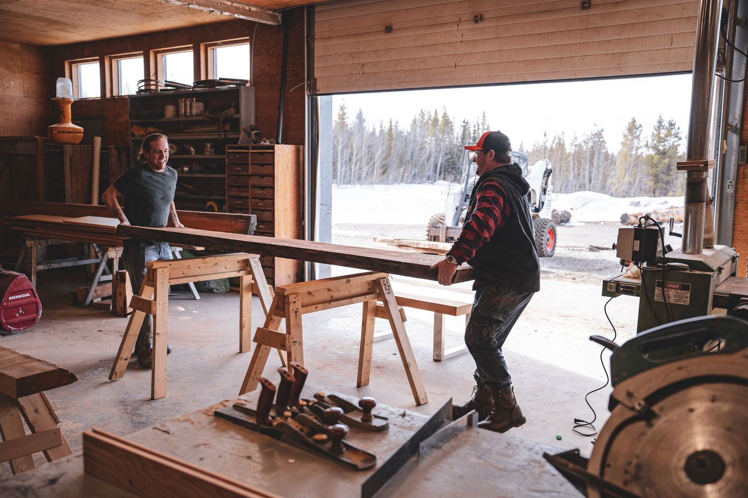 Two workers handling long wooden planks on sawhorses in an open workshop garage during Commercial Business Branding photoshoot.
