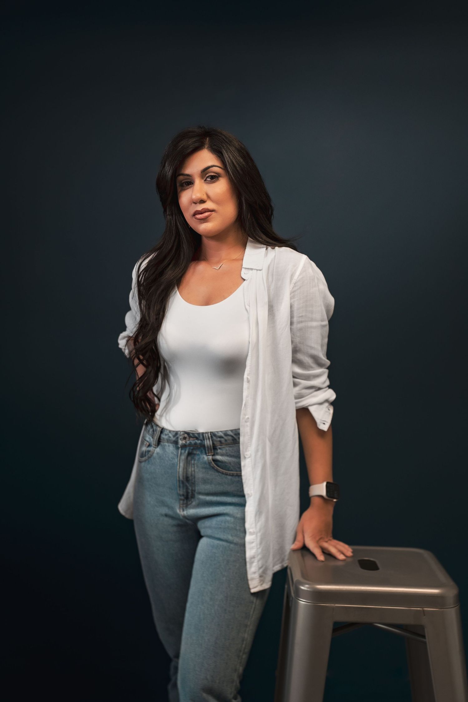Woman in white shirt and jeans leaning on metal stool against dark backdrop during her headshot photoshoot.