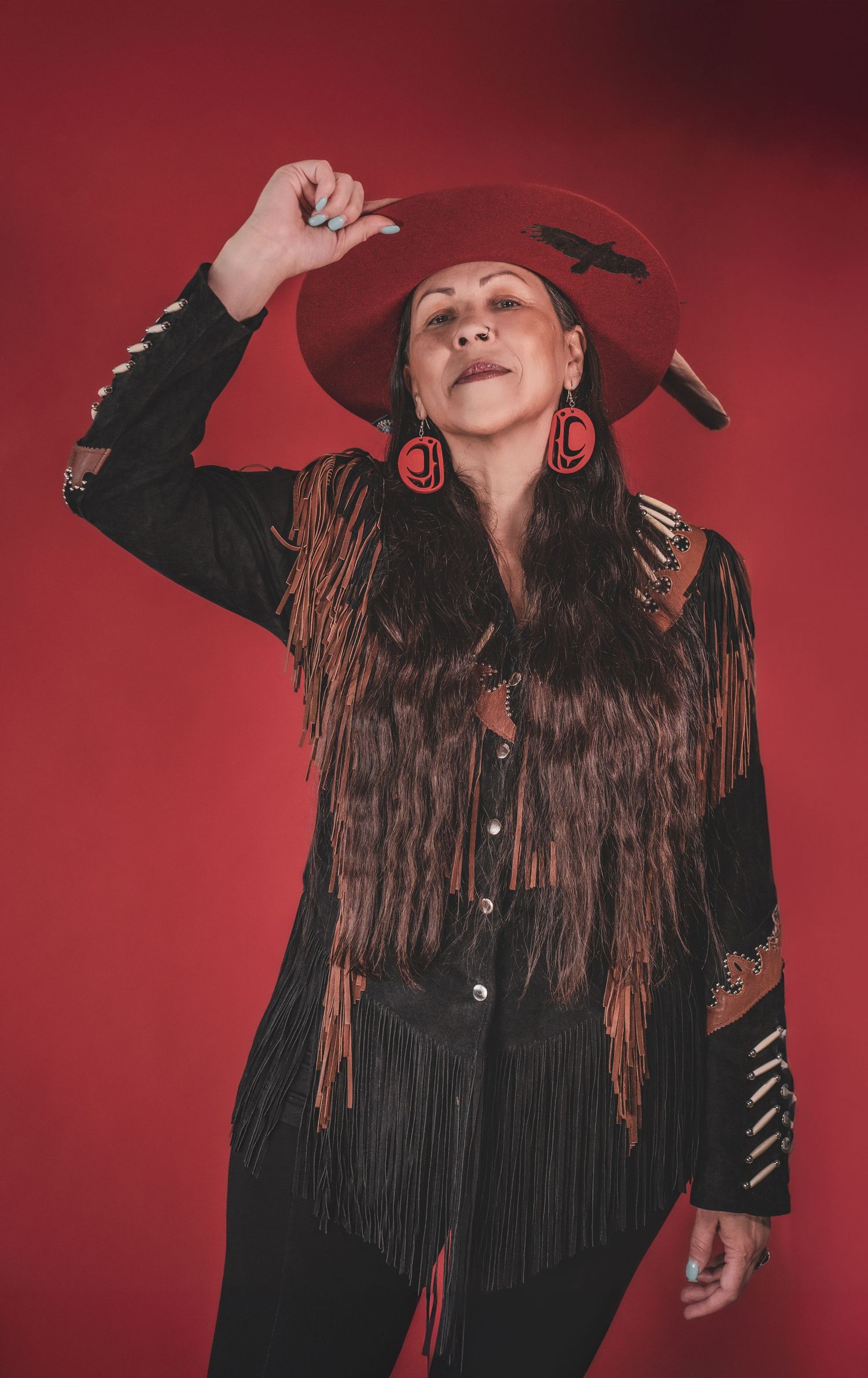 Kym Gouchie in black jacket and red wide-brim hat with red earrings posing against a red background during artist photoshoot.