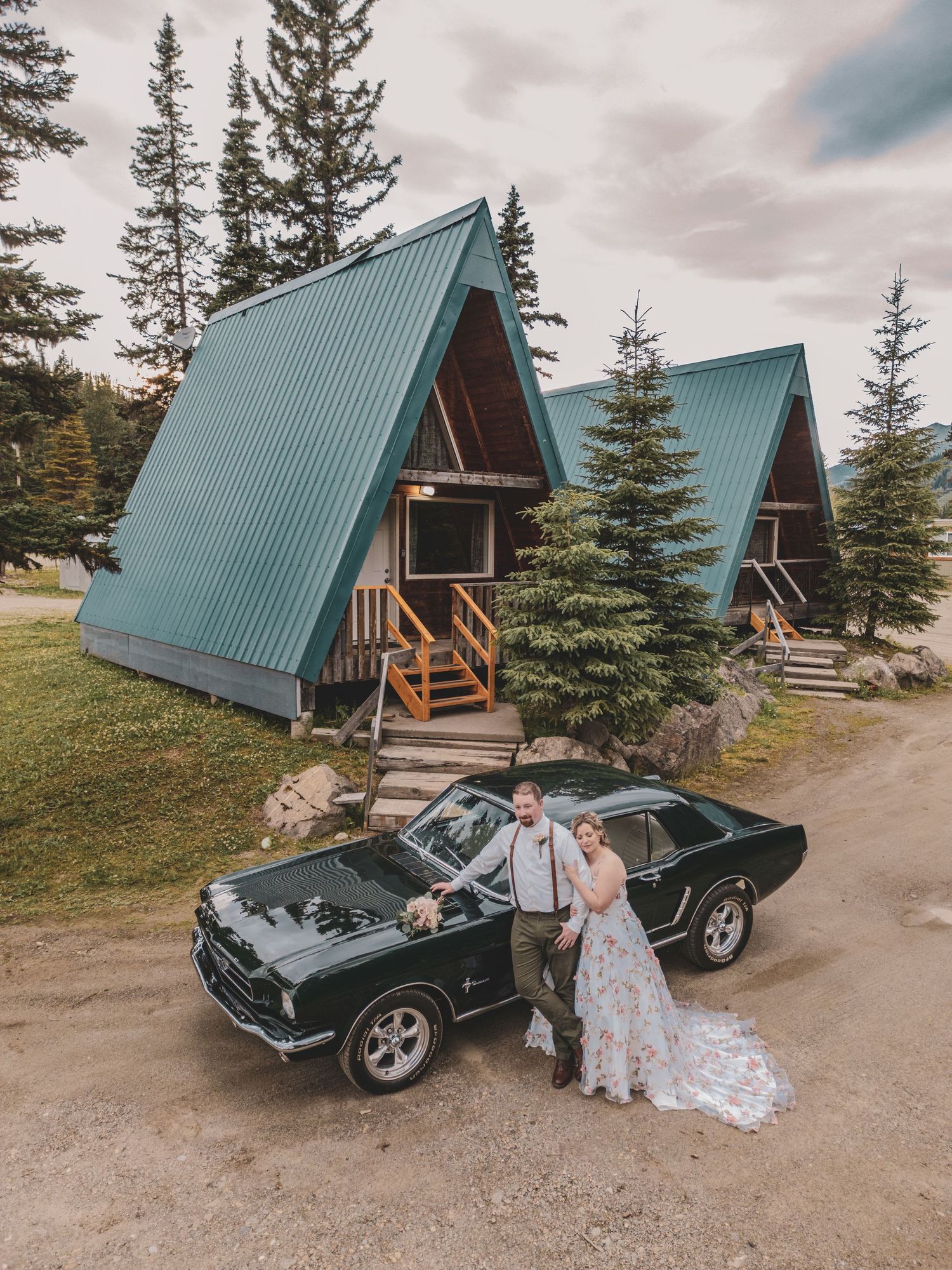 Newlyweds pose beside a vintage green Mustang in front of an A-frame cabin surrounded by pine trees.