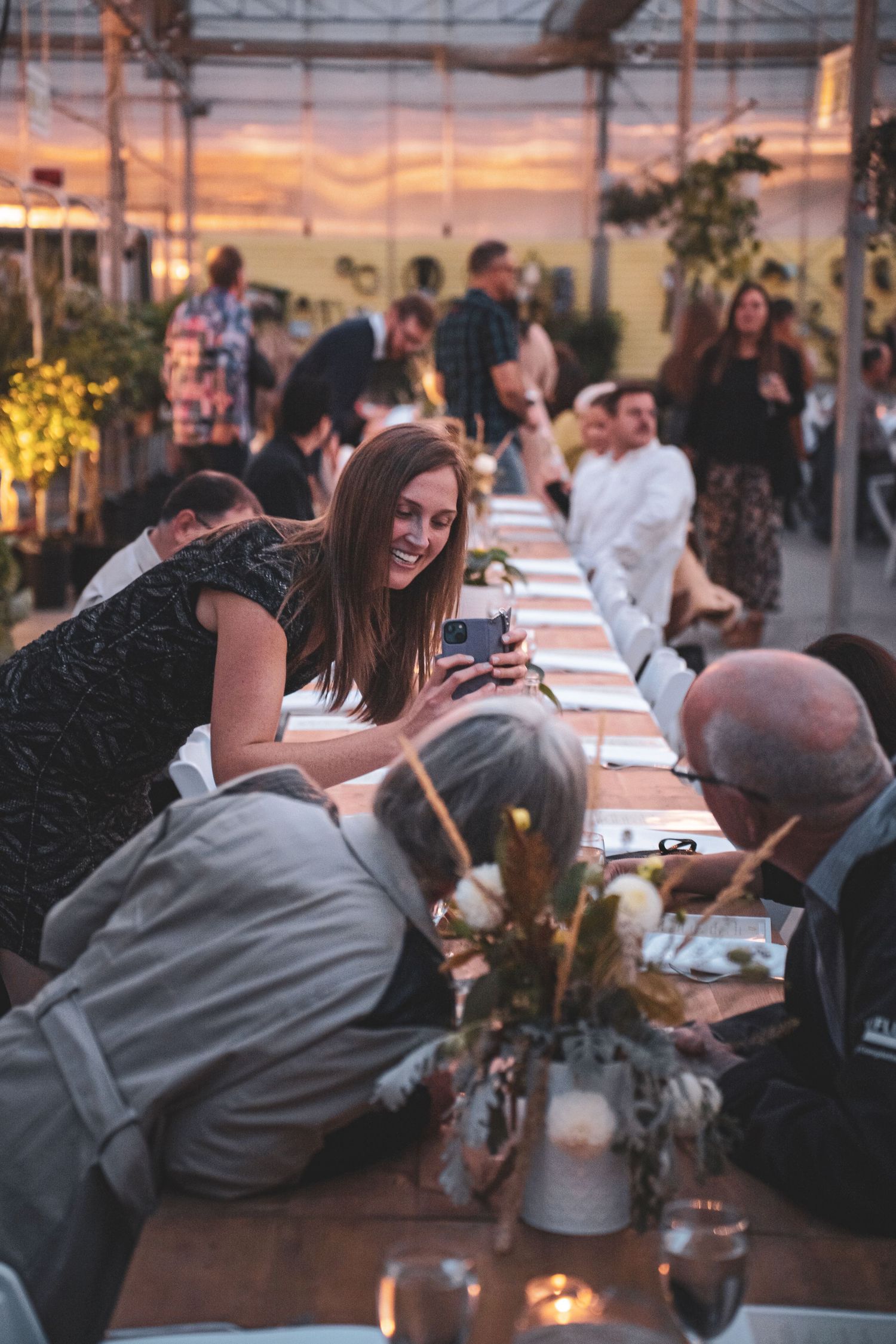 Guests enjoy a festive greenhouse dinner party along a long candlelit table decorated with flowers and greenery.