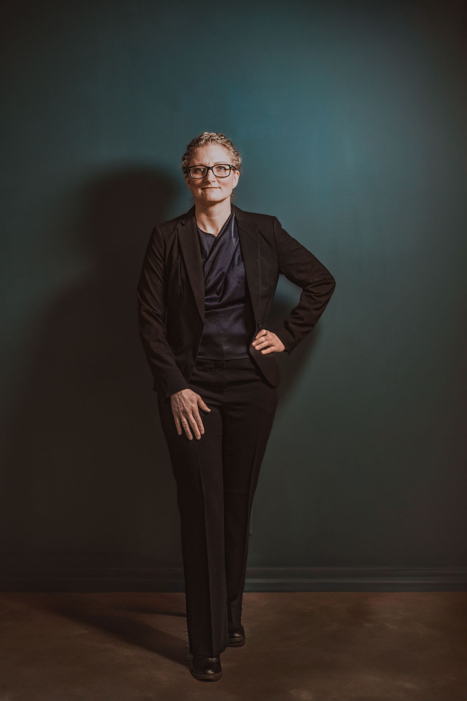 Confident woman in black suit and glasses posing against dark teal wall in full-length portrait during Commercial Business Branding photod.