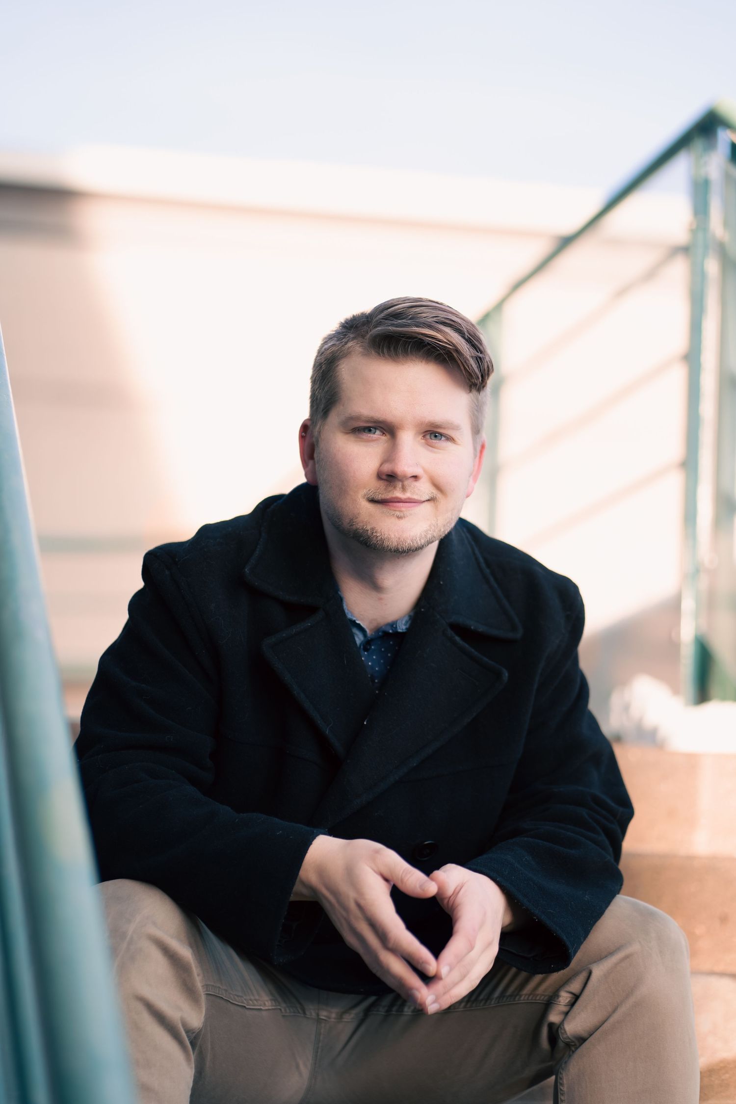 Young man in black peacoat sitting on outdoor steps with hands clasped, smiling at camera during Commercial headshots.