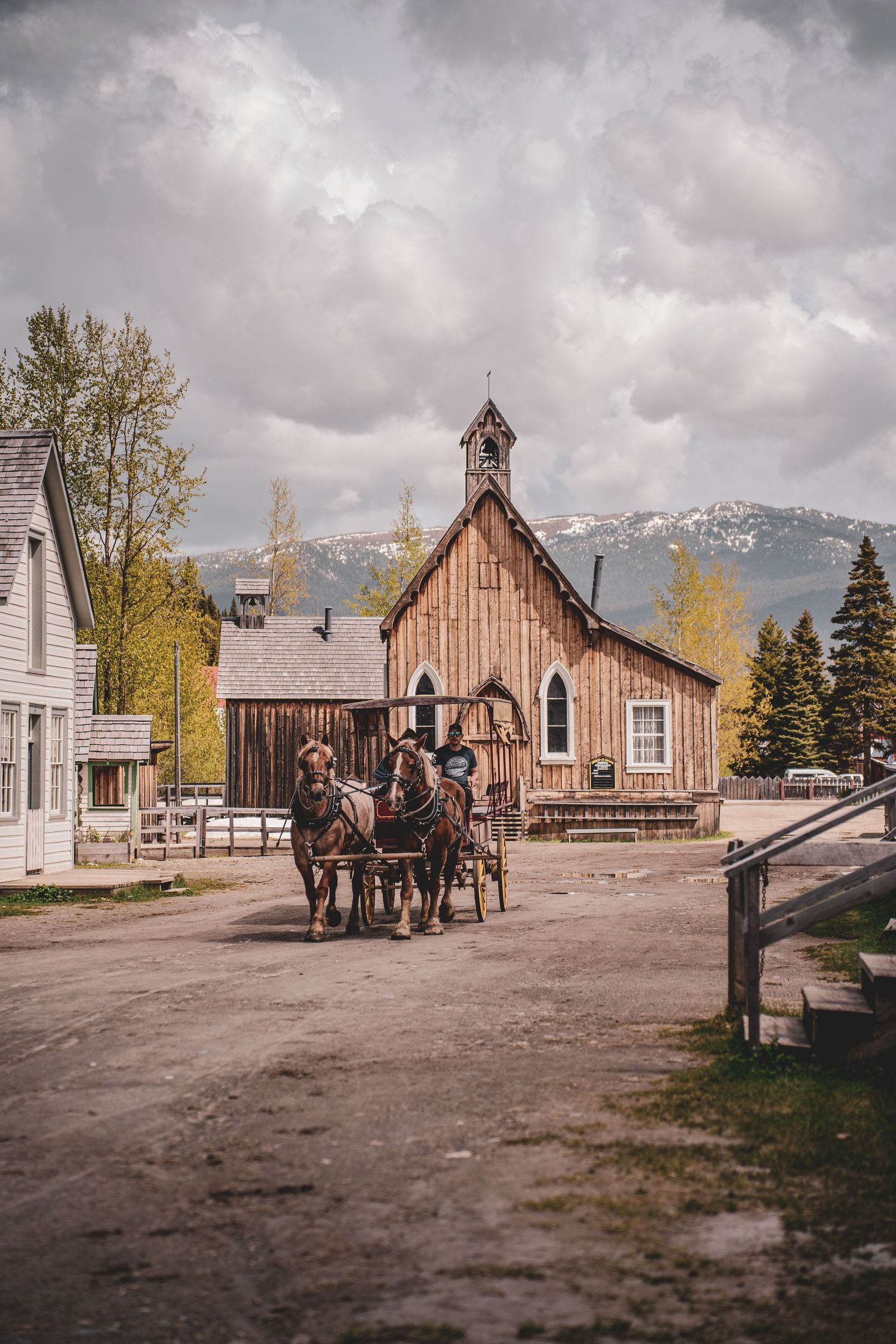 Horse-drawn carriage passing a rustic wooden church in a historic western town with mountain backdrop.