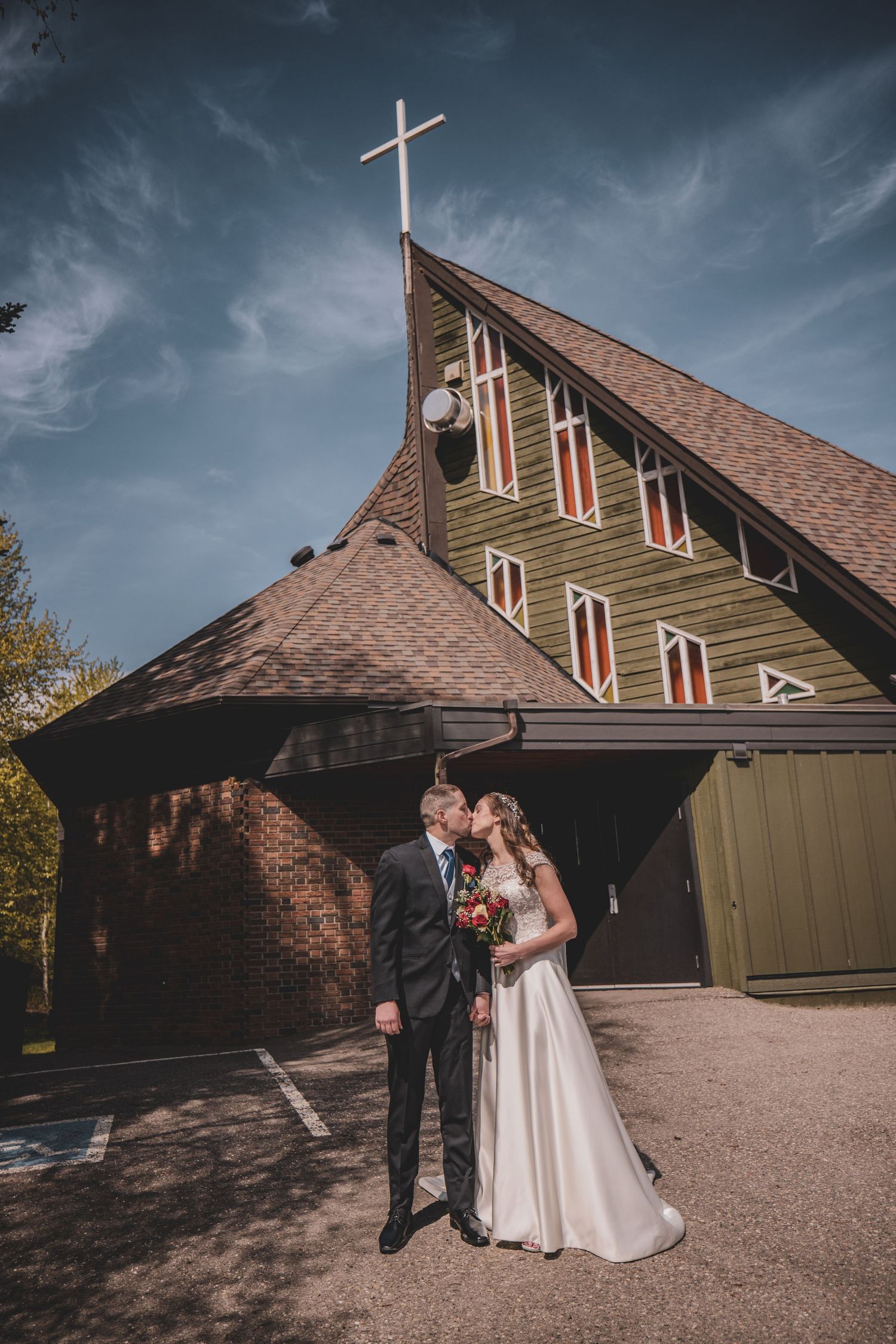 Bride and groom share a kiss outside a charming A-frame church with a cross on a sunny day, at a Prince George BC church venue.
