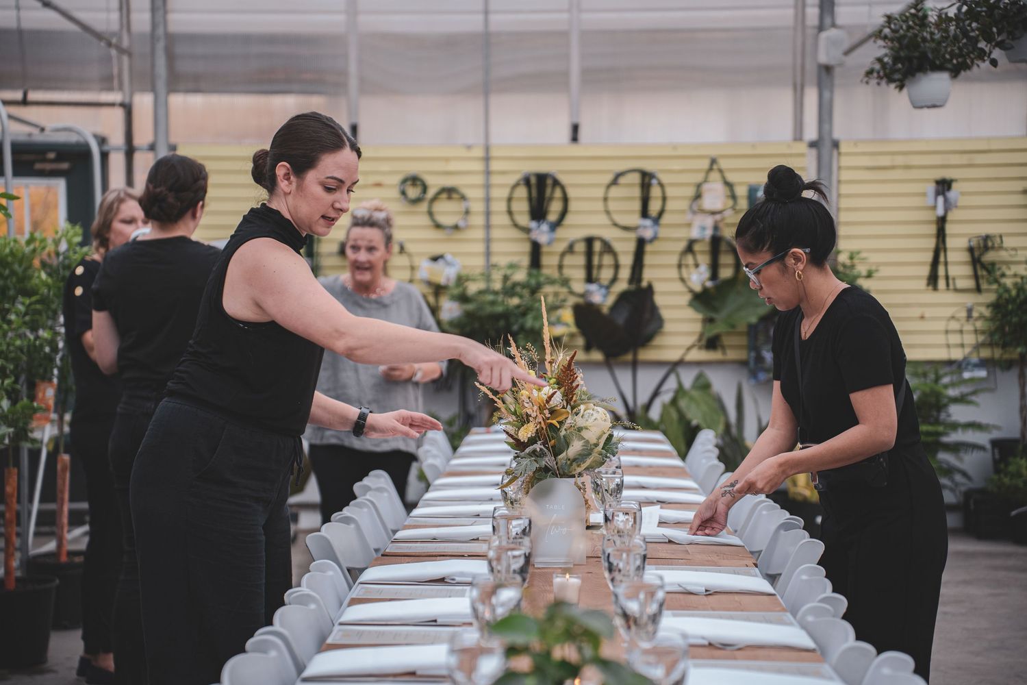 Two women in black attire arranging floral centerpieces on a long elegantly set dining table.
