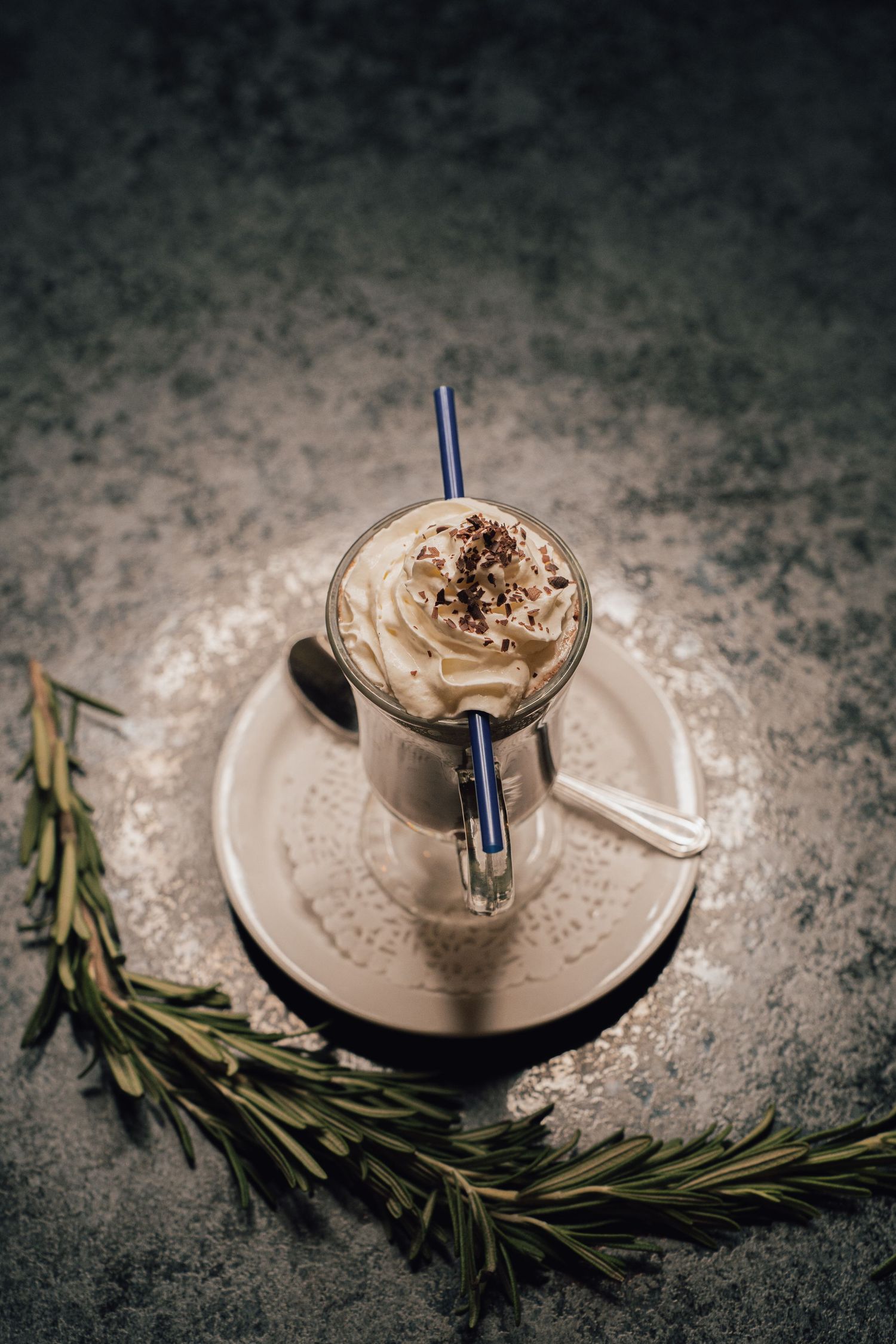 Whipped cream topped coffee drink with blue straw on saucer surrounded by fresh rosemary sprigs.