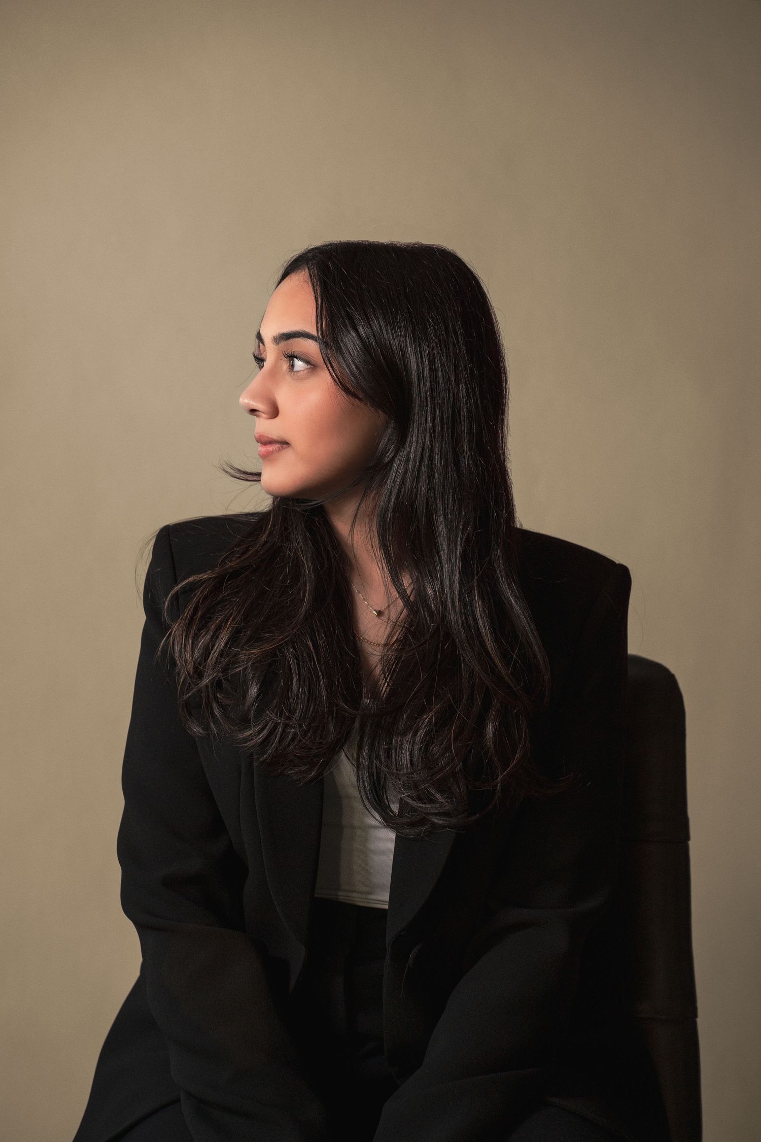 Young woman in black blazer seated looking sideways against neutral background during her real estate headshot photoshoot.