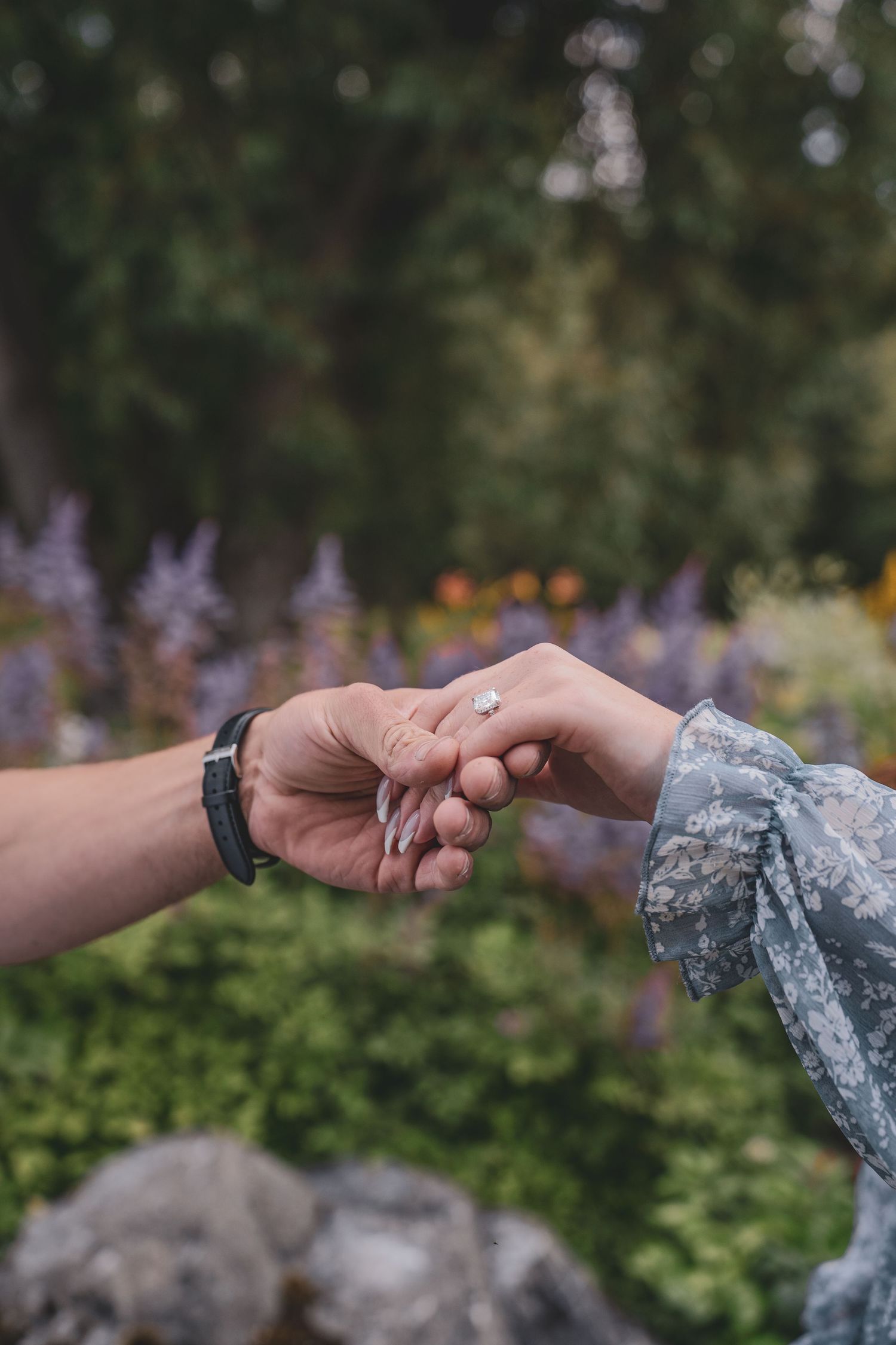 Close-up of couple holding hands with engagement ring in a colorful garden setting.