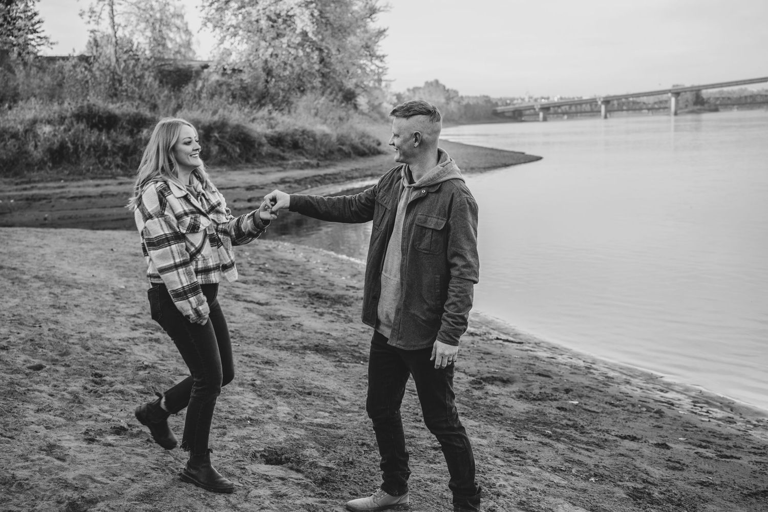 Black and white photo of couple holding hands and laughing on a riverside beach.