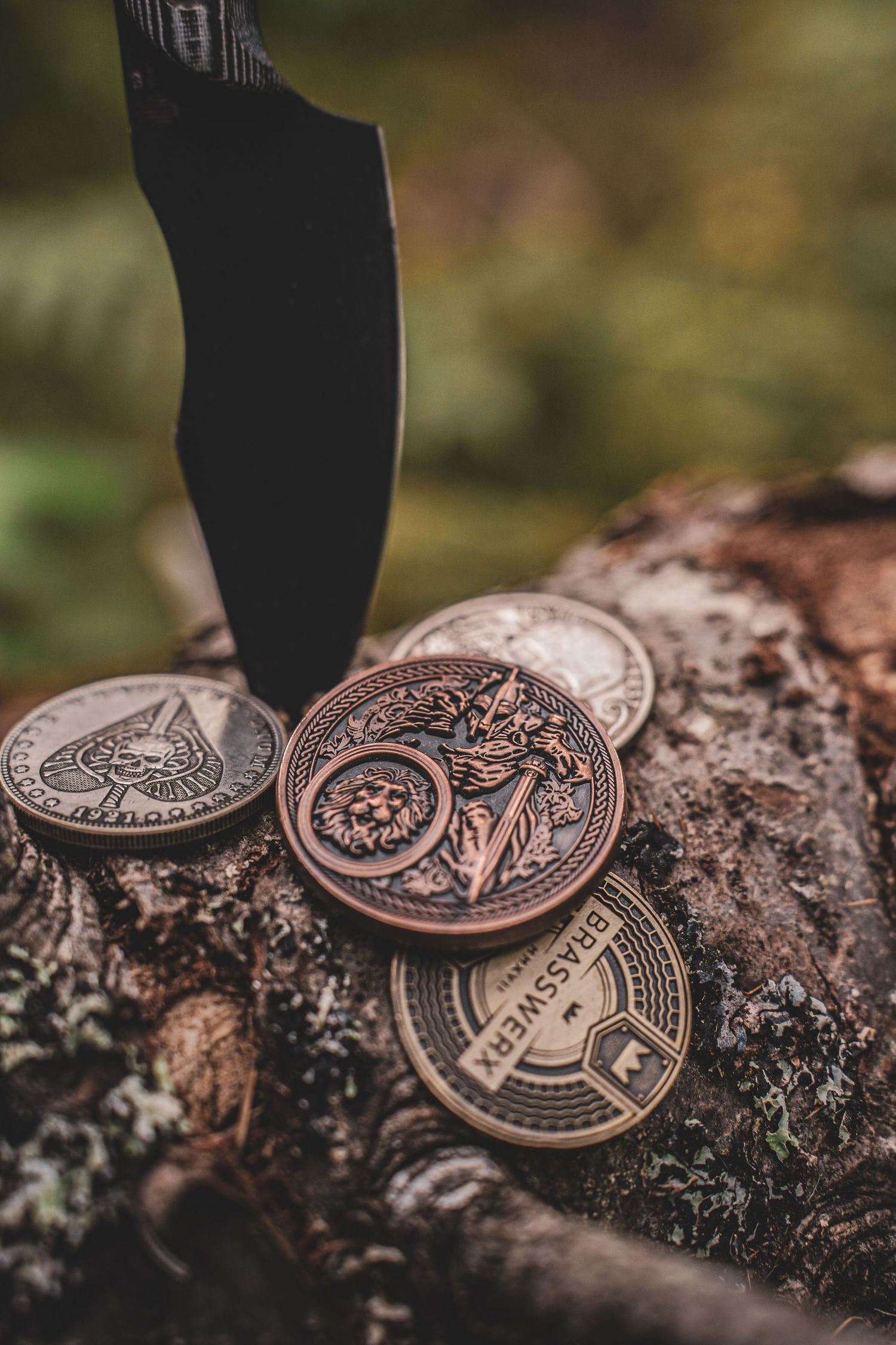 Product photography of Black tactical knife stabbed into mossy log surrounded by decorative brass challenge coins.