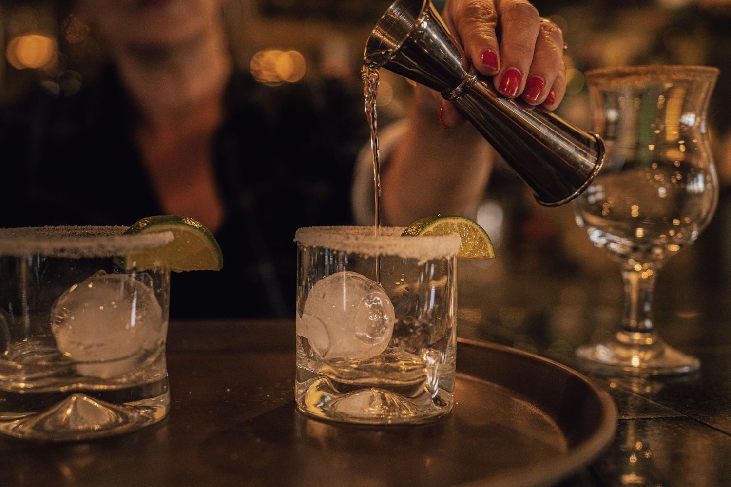 Bartender pouring cocktail into ice-filled glasses garnished with lime on a bar tray.