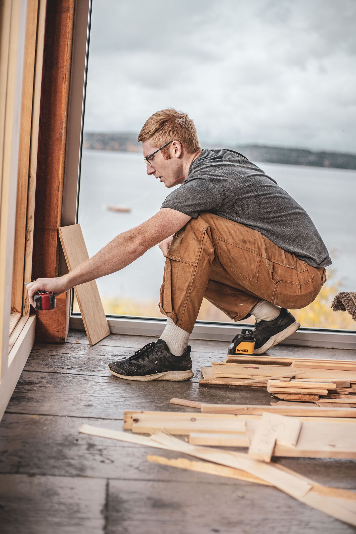 Young carpenter in safety glasses measuring wood trim near large windows with waterfront view.