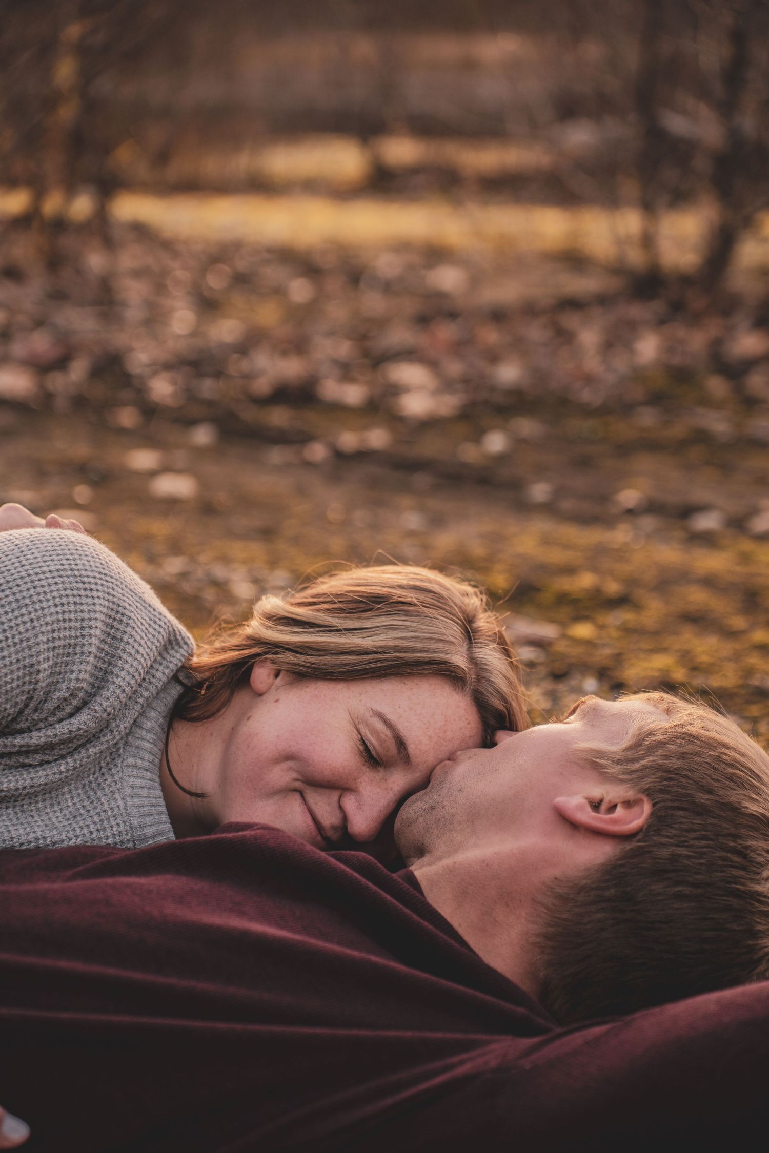 Couple sharing an intimate almost-kiss while lying on a burgundy blanket in an outdoor autumn setting.