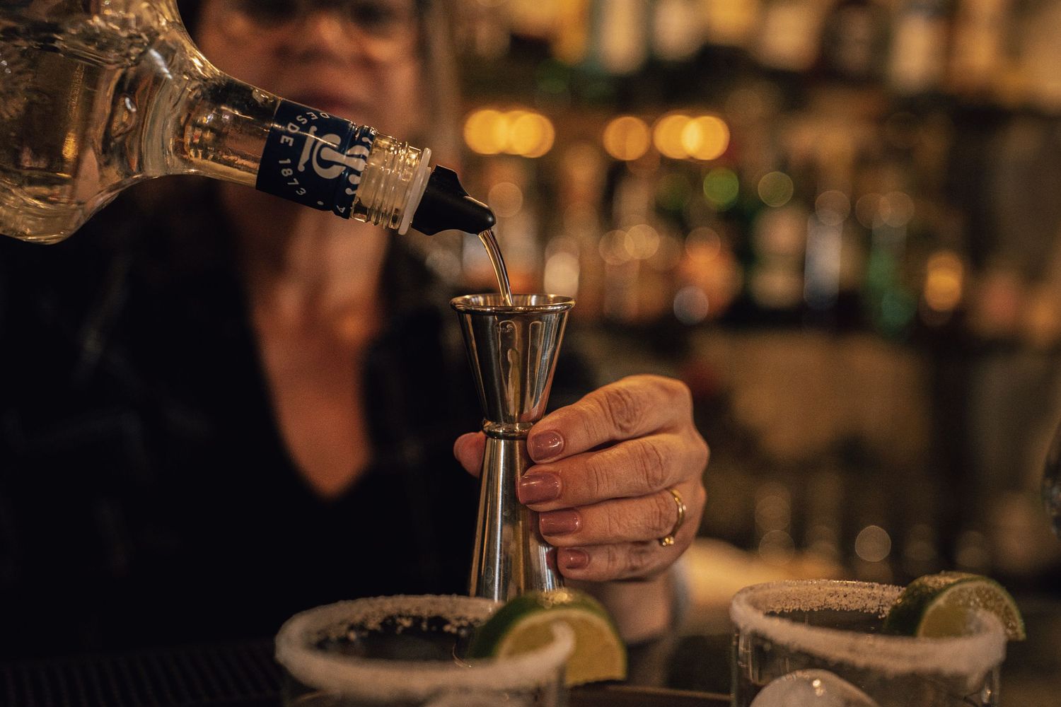 Bartender measuring spirits into a jigger for a margarita with salted rim and lime garnish.