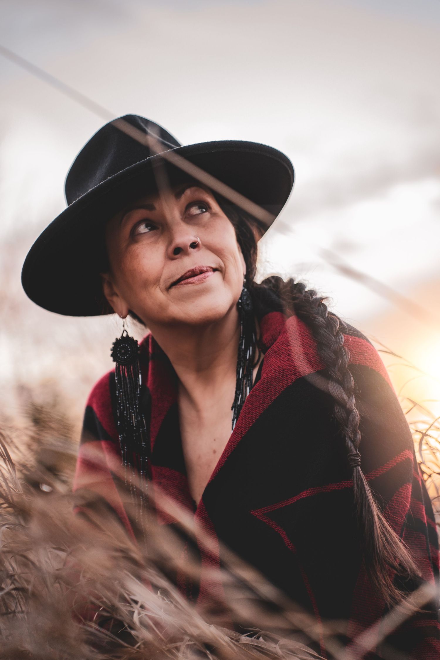 Kym Gouchie in black wide-brim hat and red blanket wrap sitting in golden grass at sunset during Commercial Business Branding photoshoot.