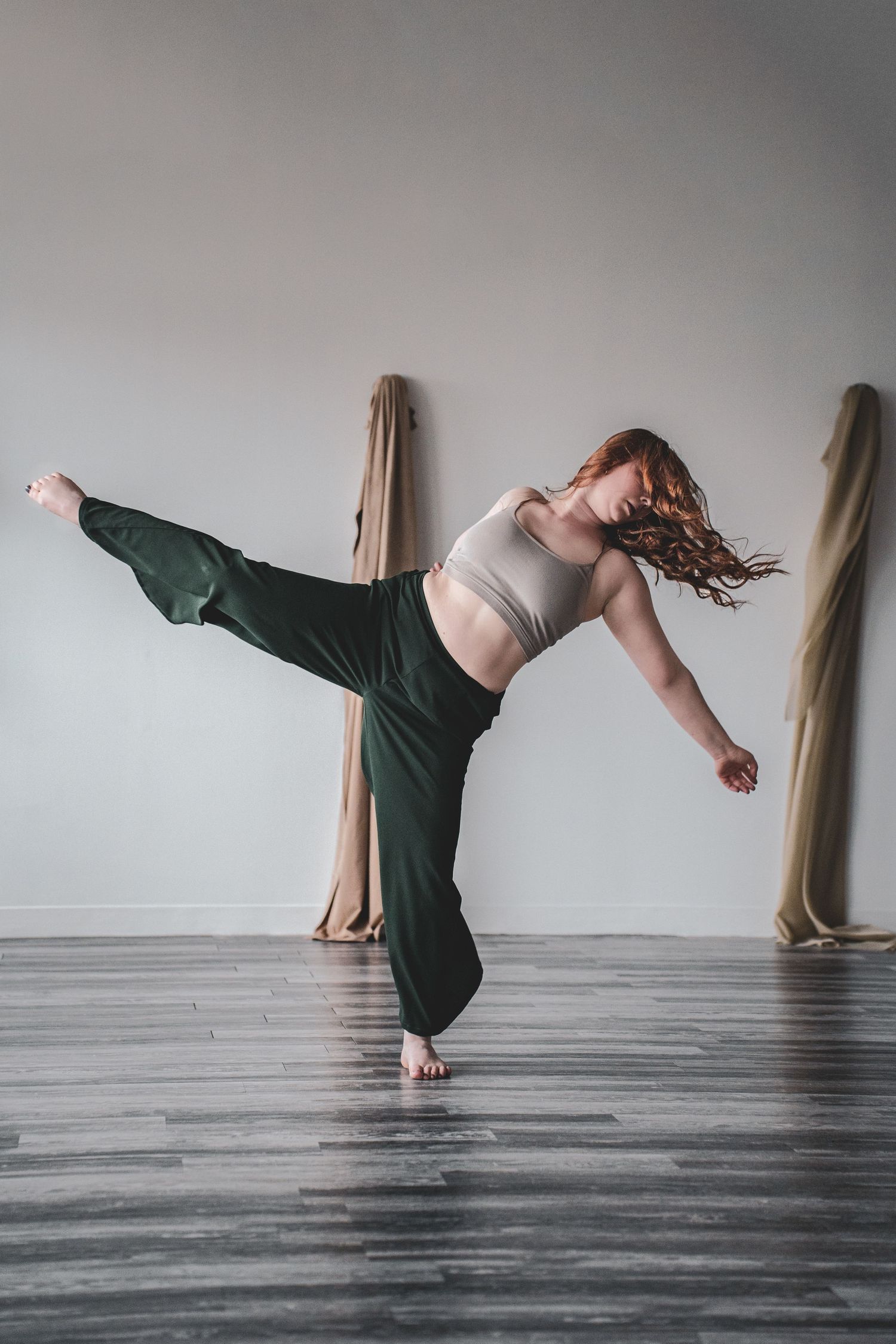 A female dancer balances on one foot with leg extended high, wearing green wide-leg pants in a dance studio, during branding photoshoot.