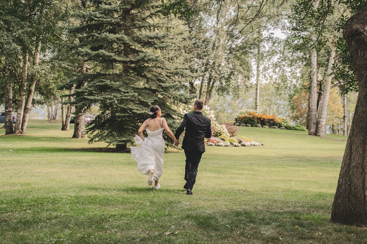 Bride and groom holding hands running through a lush green park surrounded by tall trees.