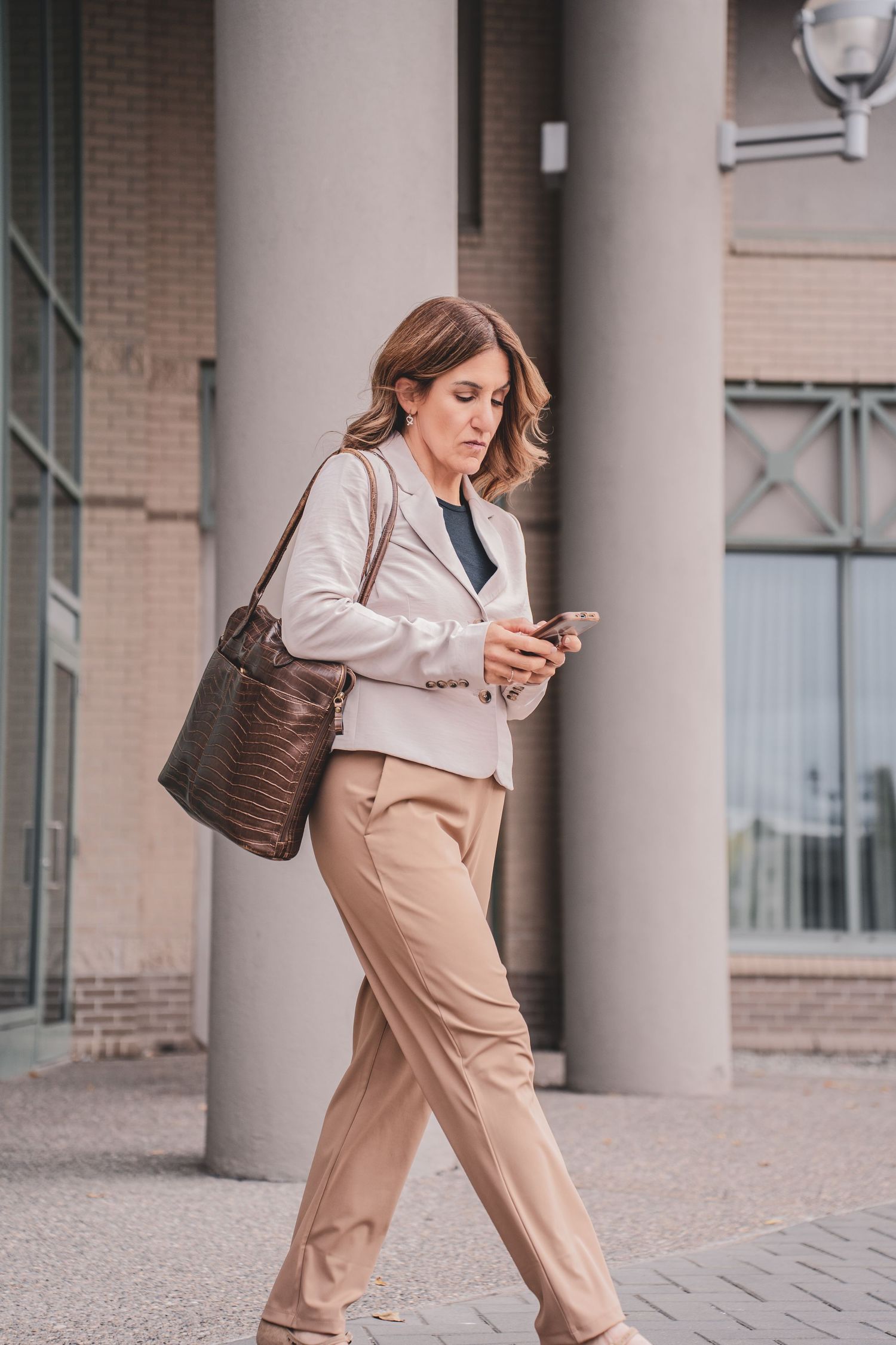 Professional woman in beige blazer checking smartphone while walking past office building.