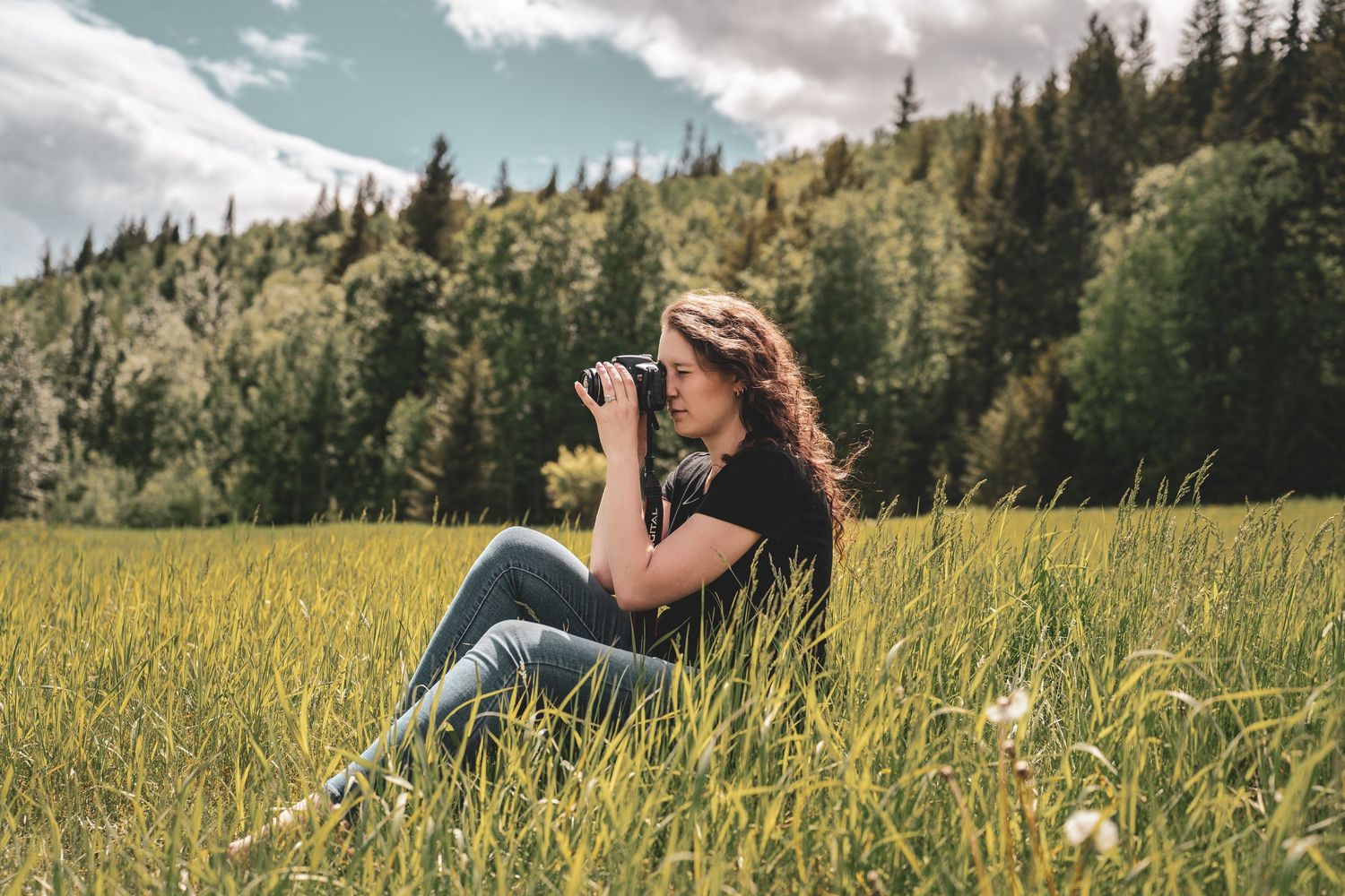 A woman with long brown hair sits in a green meadow photographing with a DSLR camera outdoors for photographer portfolio shoot.