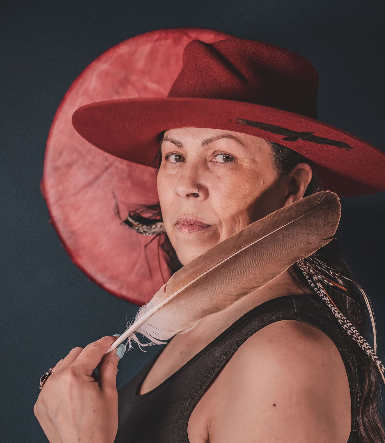 Kym Gouchie in in red wide-brim hat holding a large feather, looking forward against dark background during musician photoshoot.