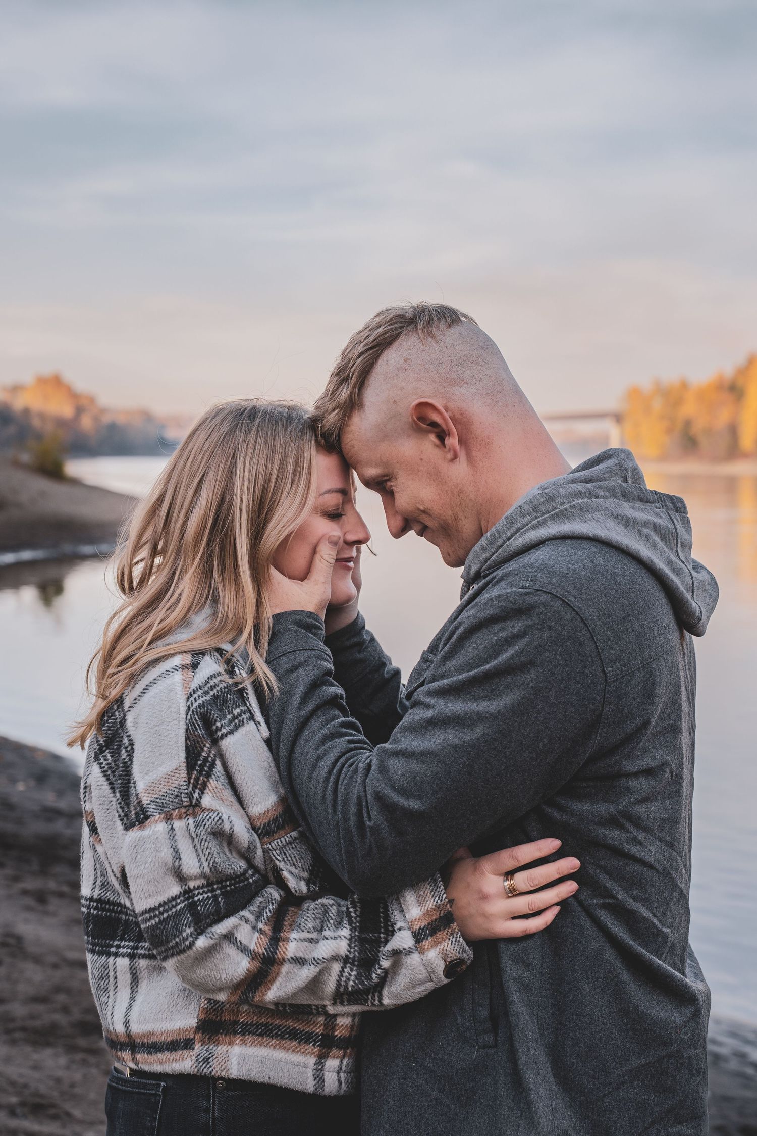 Romantic couple touching foreheads by a calm river during a golden autumn sunset.