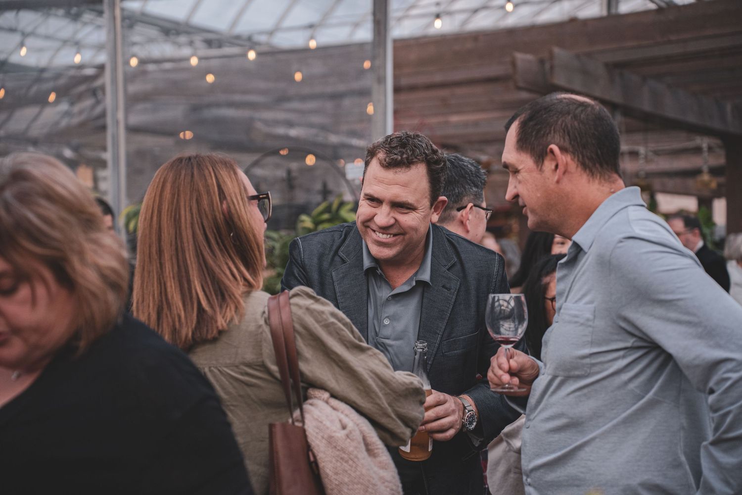 People socializing at an indoor greenhouse event with string lights, enjoying wine and conversation.