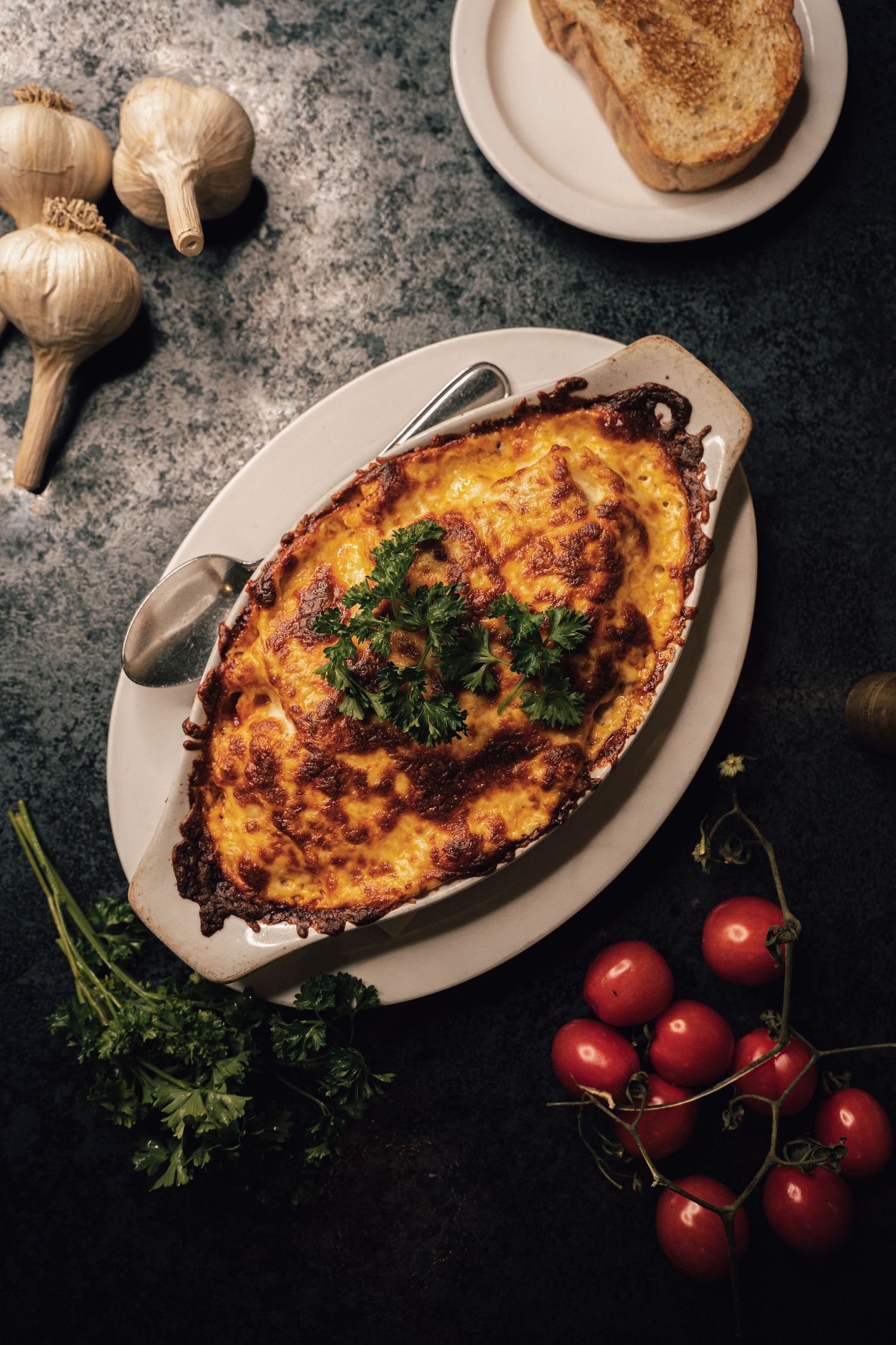 Golden-baked moussaka garnished with fresh parsley in a white oval dish, served with cherry tomatoes and garlic.