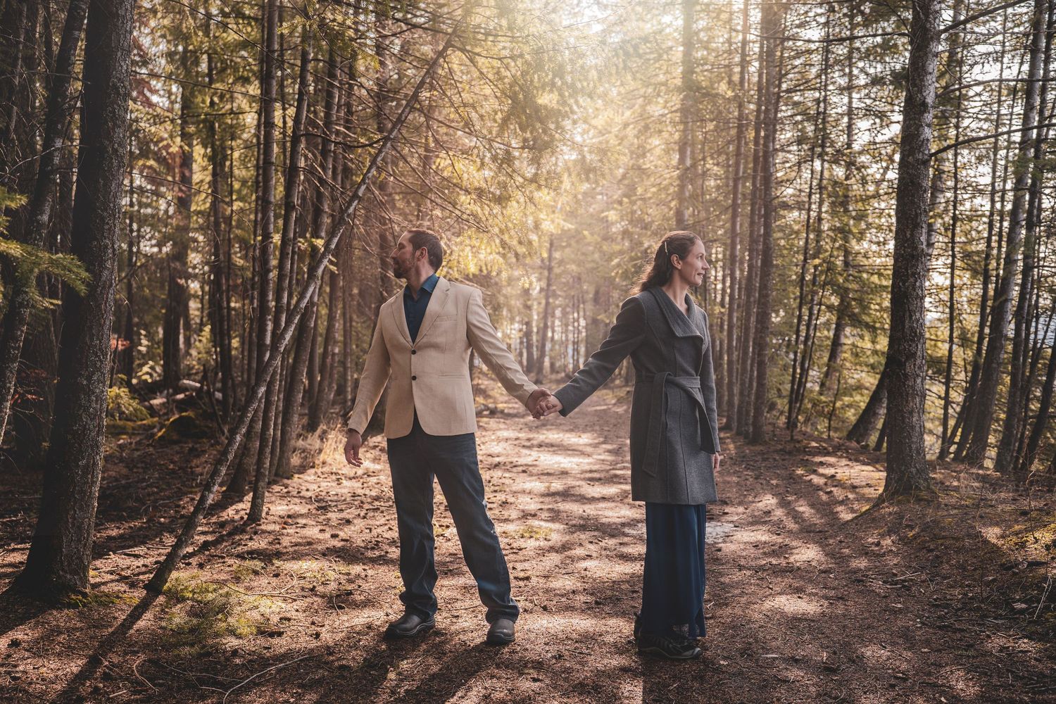 Couple holding hands back-to-back on a sunlit forest path surrounded by tall pine trees.