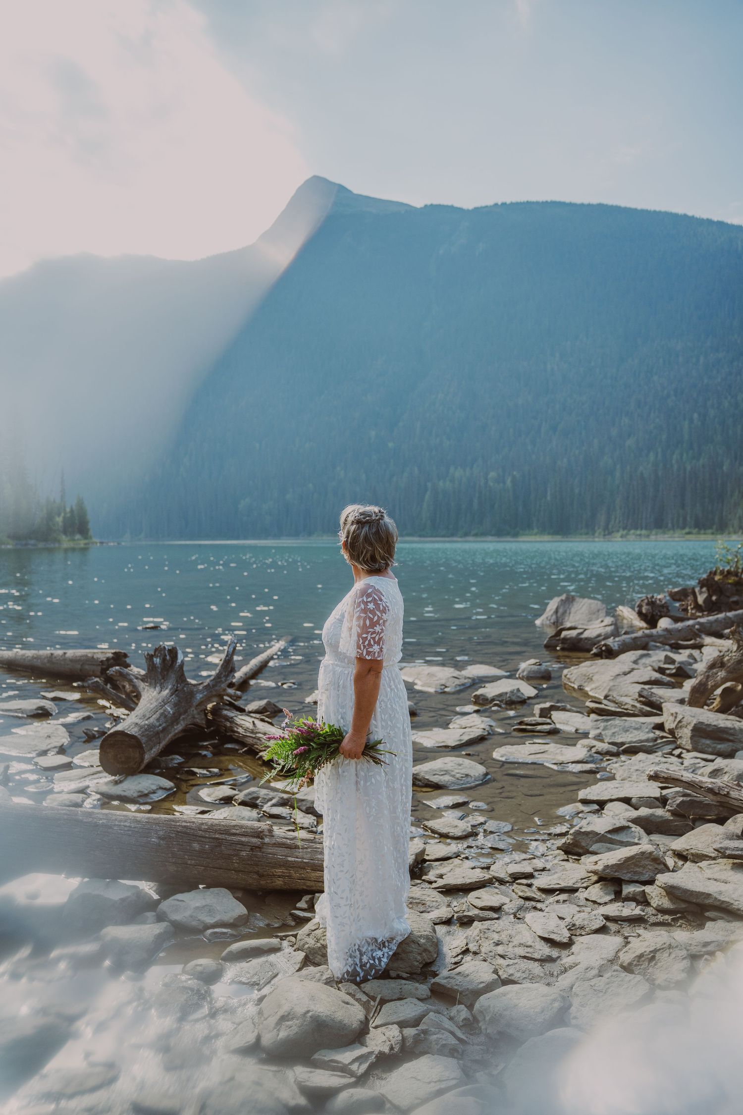 Bride in elegant white lace dress standing on rocky lakeside with majestic mountain backdrop glowing.