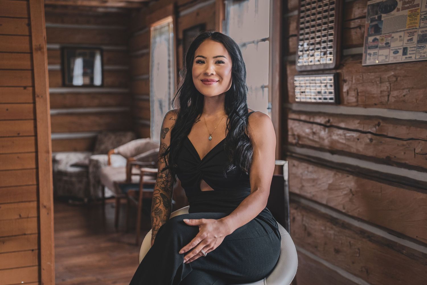 Smiling woman with tattoos in black dress sitting in rustic log cabin interior.