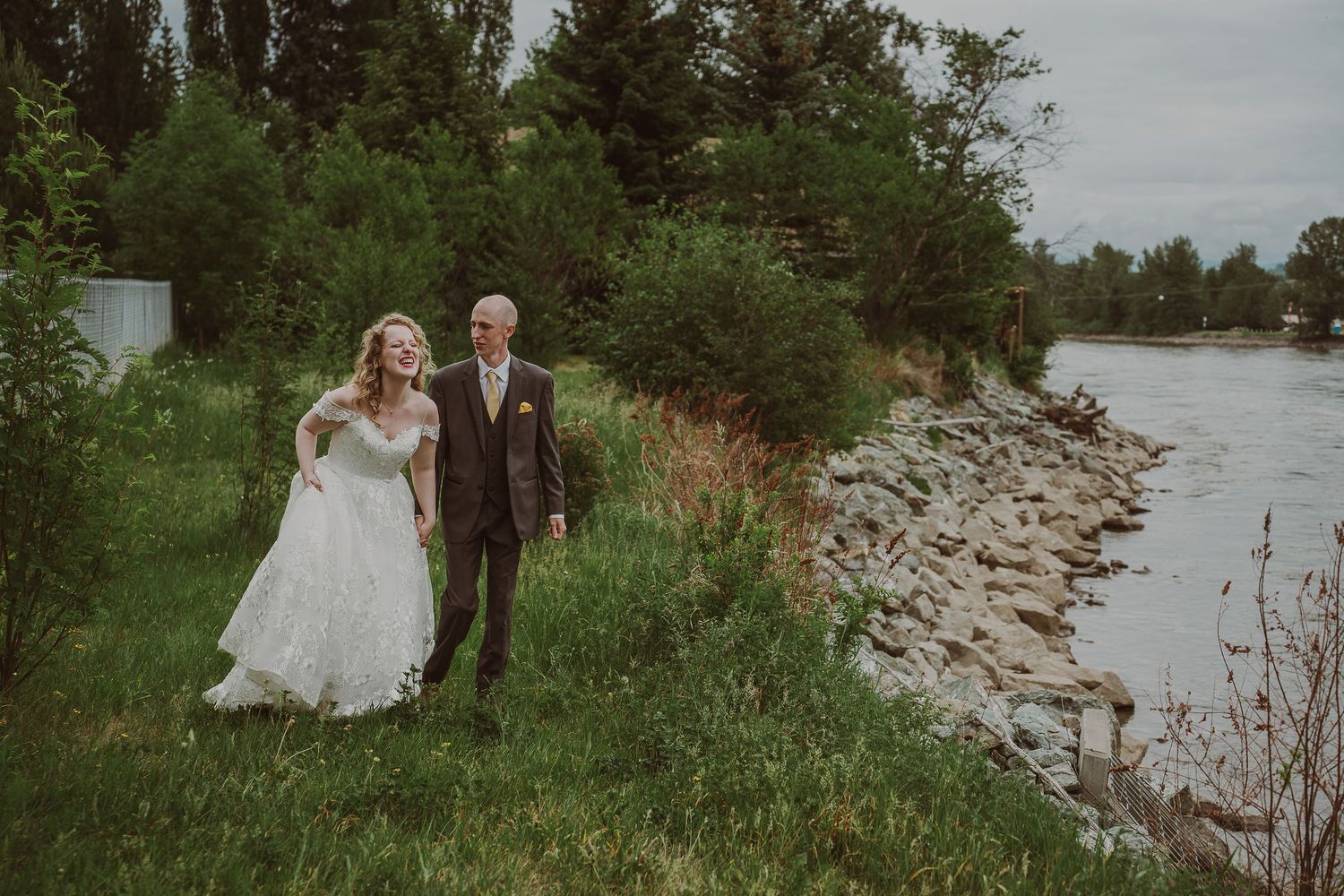 Smiling bride and groom walking hand in hand along rocky riverbank of the Northern Lights Winery for their photoshoot with Free West Photo.