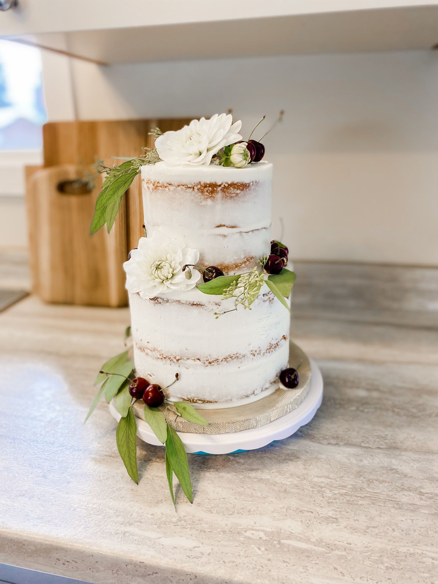 Three-tier semi-naked wedding cake decorated with white flowers, cherries, and greenery on marble stand offered by Prince george cake maker.