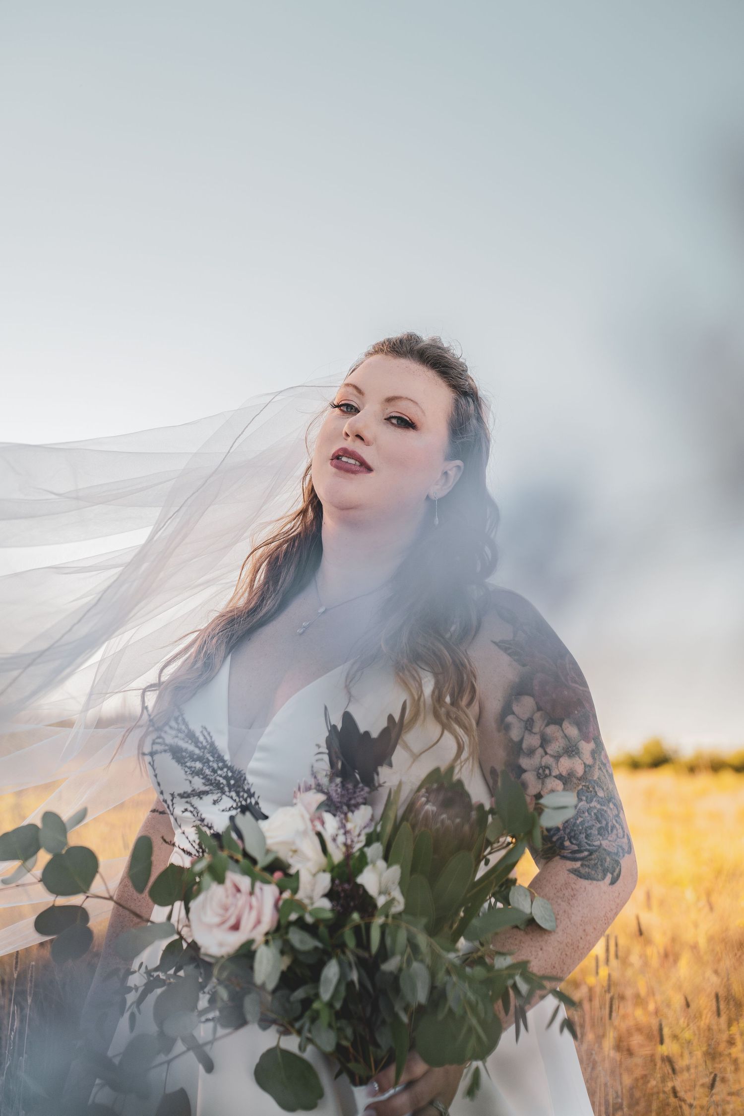 Bride in white gown with tattoos holds bouquet as veil flows in wind against golden field backdrop at her country wedding day.