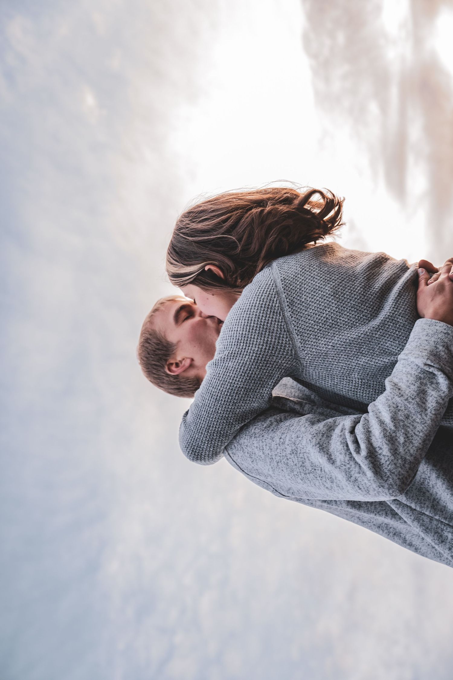 Romantic couple sharing a kiss outdoors in winter, man lifting woman against a snowy sky backdrop.