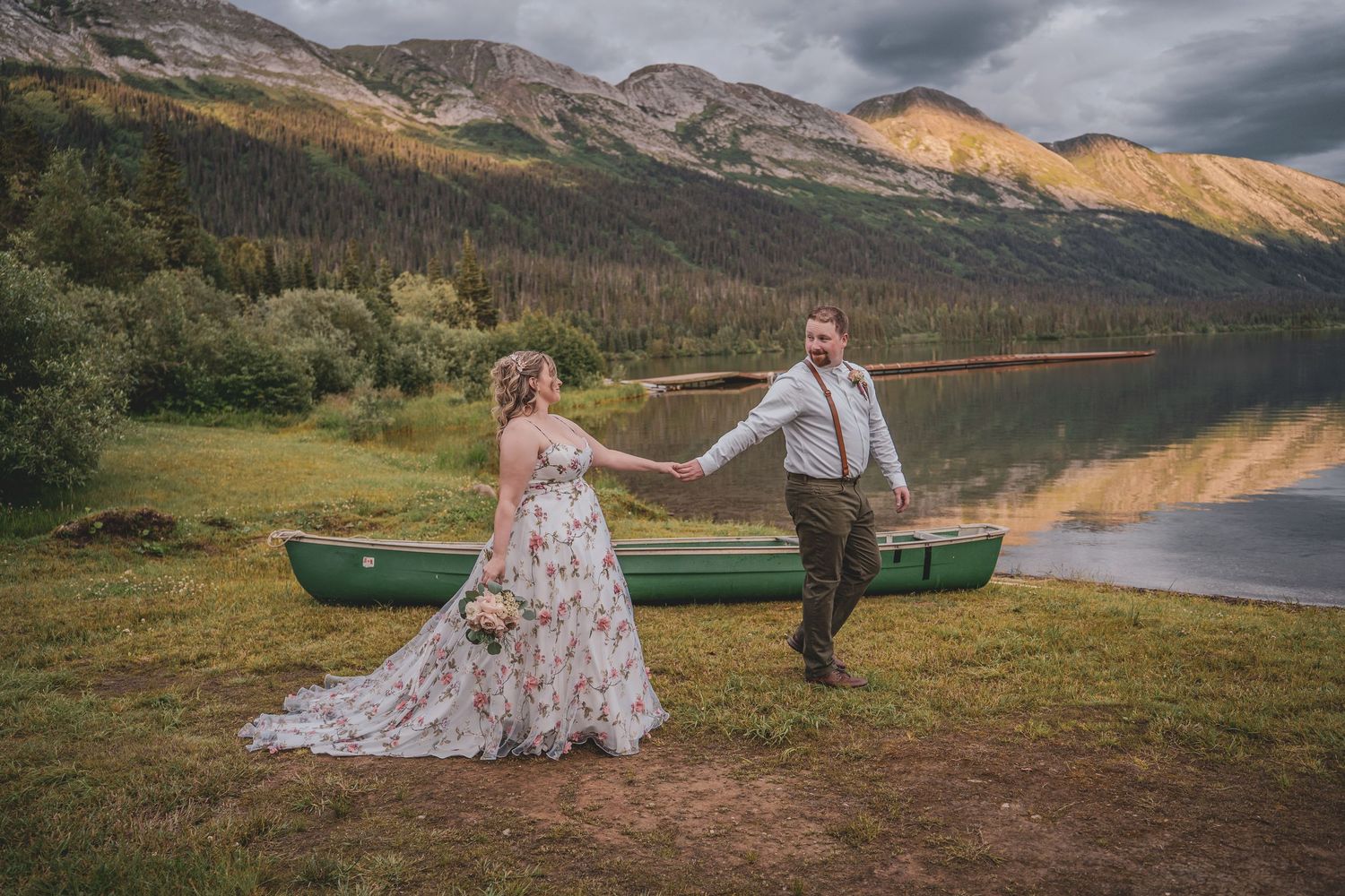 Bride and groom hold hands near a green canoe by a mountain lake during their outdoor elopement ceremony.