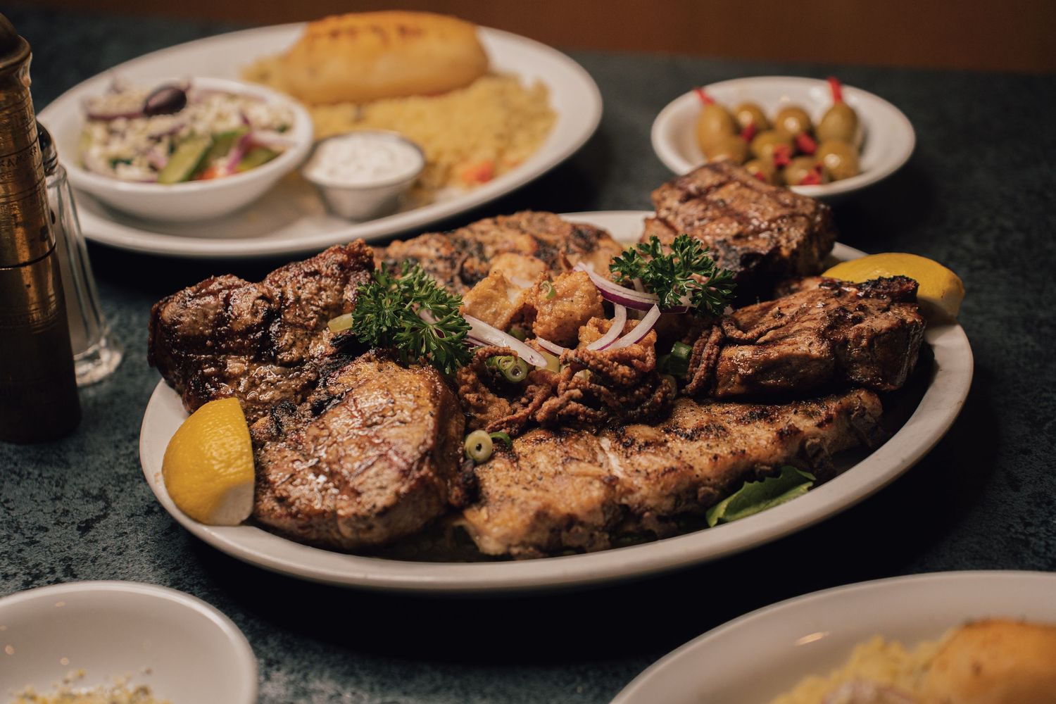 Close-up of a grilled mixed meat platter with lemon, parsley, and side dishes in background.