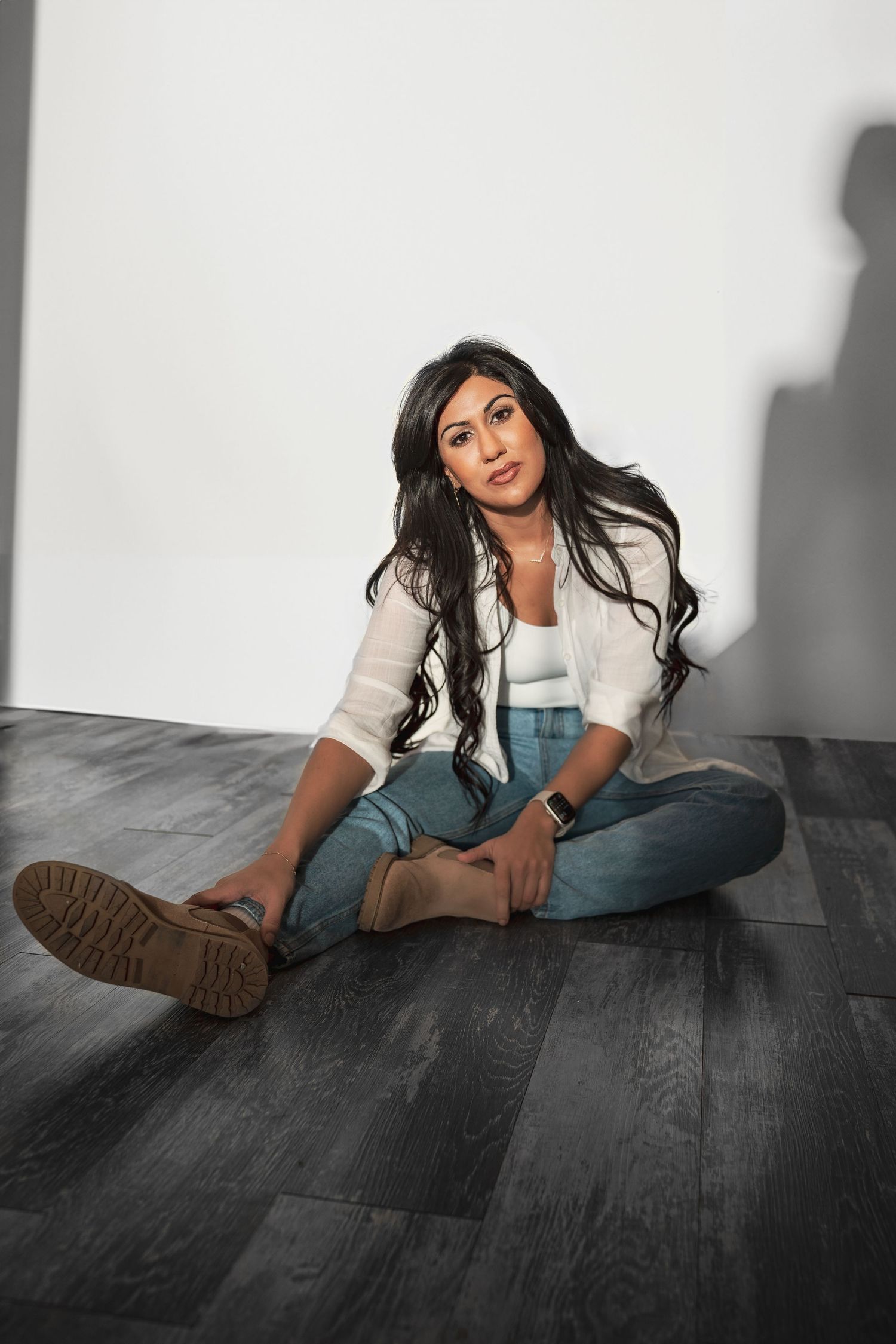 A woman with long dark hair sits cross-legged on a dark hardwood floor wearing jeans and a white shirt during her business photoshoot.