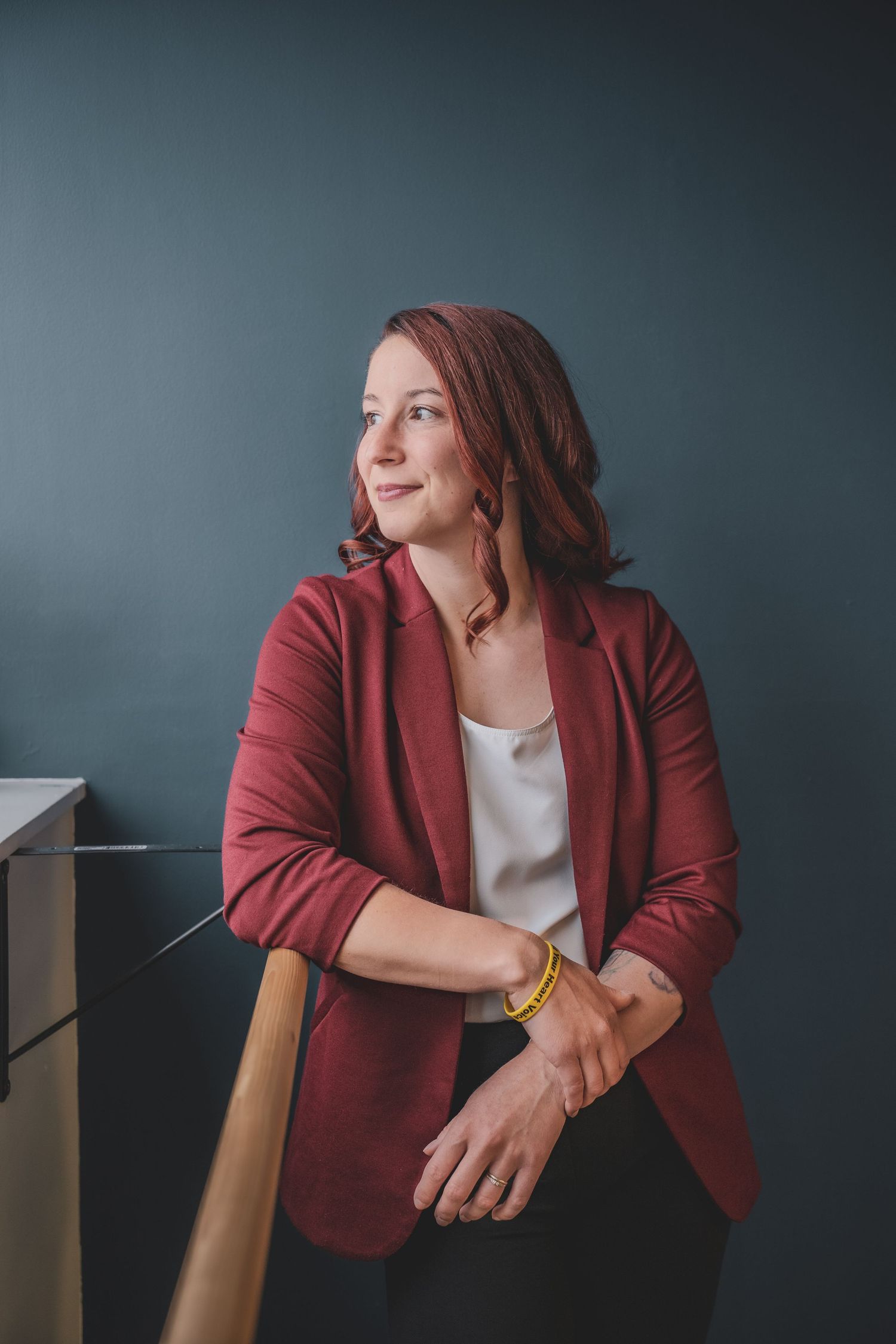 Woman in red blazer leaning on railing against teal wall looking away thoughtfully during her commercial branding photoshoot.