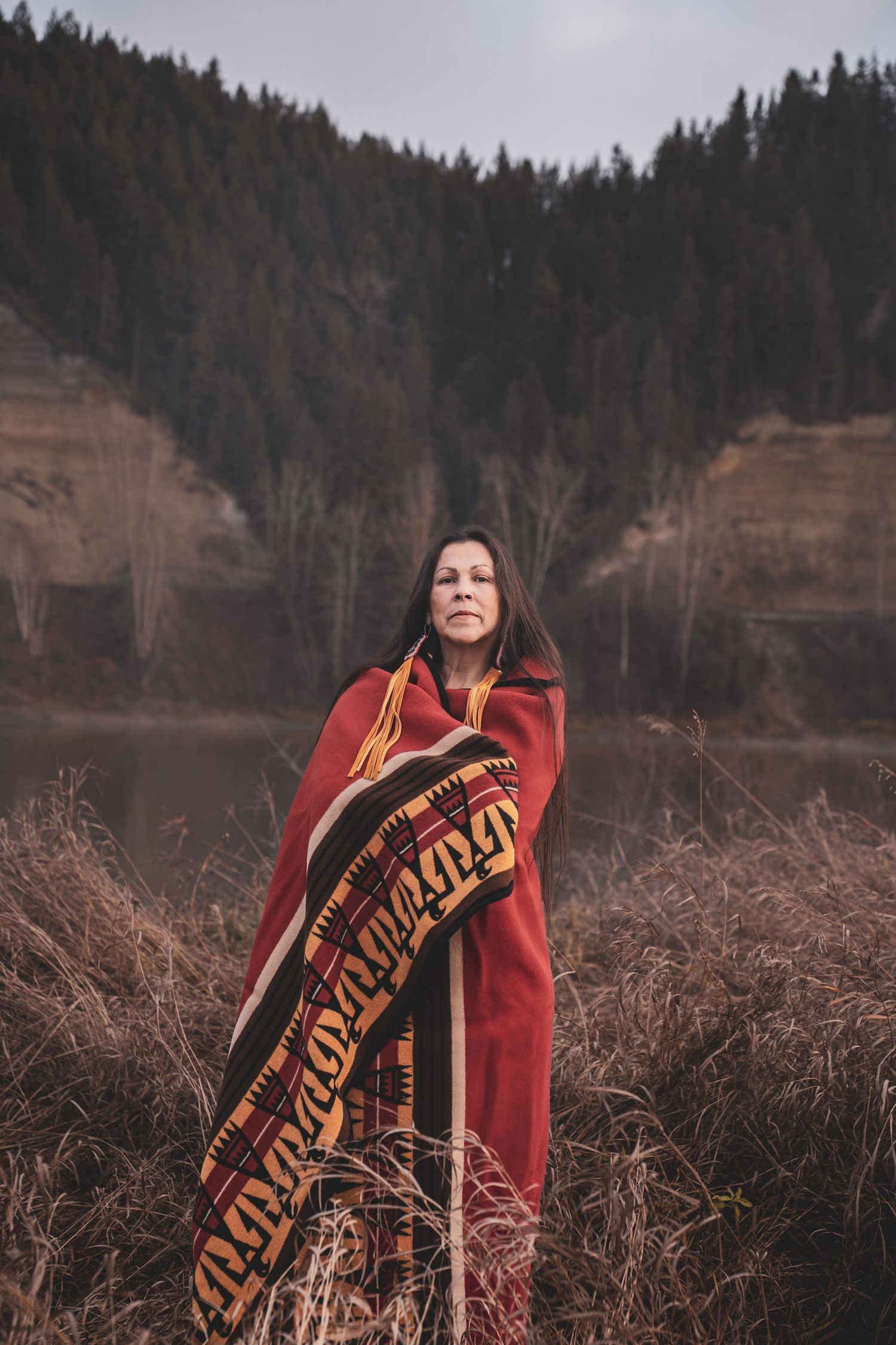 Kym Gouchie wrapped in red Indigenous patterned blanket standing in tall grass near river with forest backdrop during portrait photoshoot.